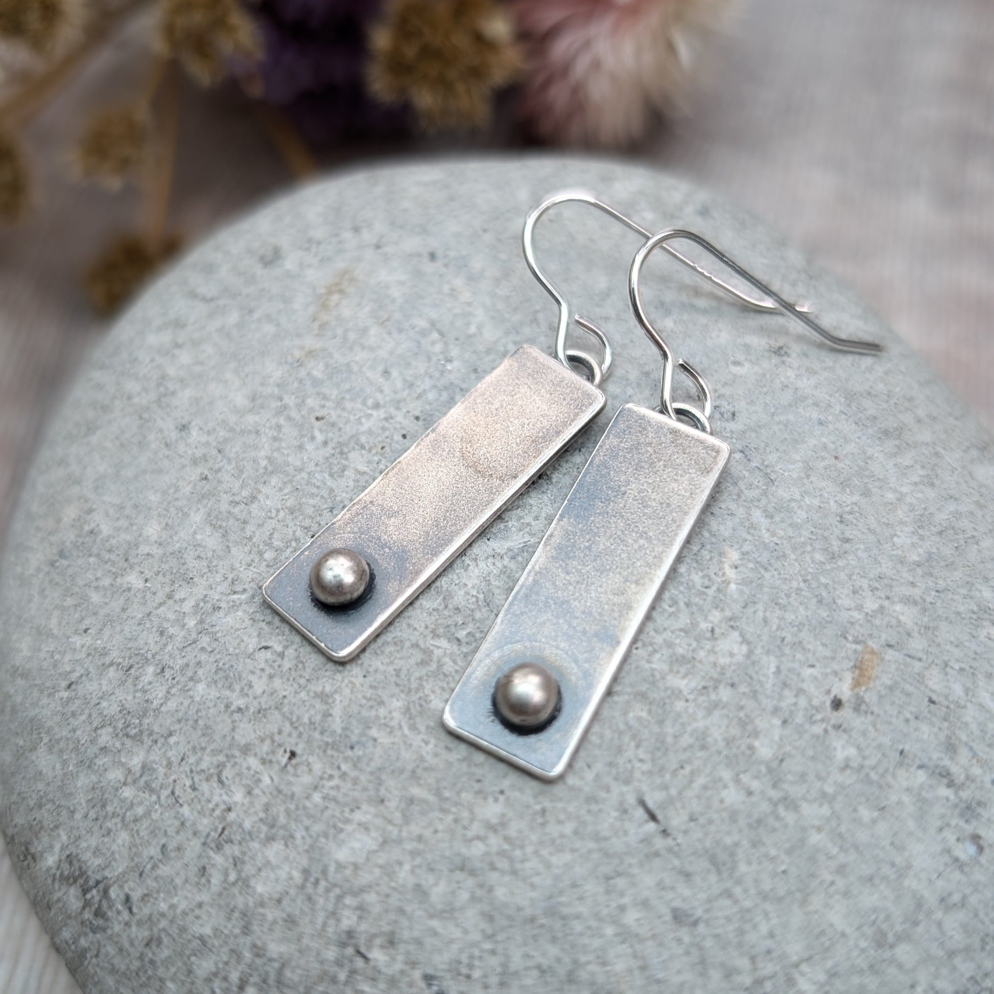 A pair of rectangular oxidized silver earrings with a small round silver pebble at the bottom of each rectangle, placed on a smooth gray stone.