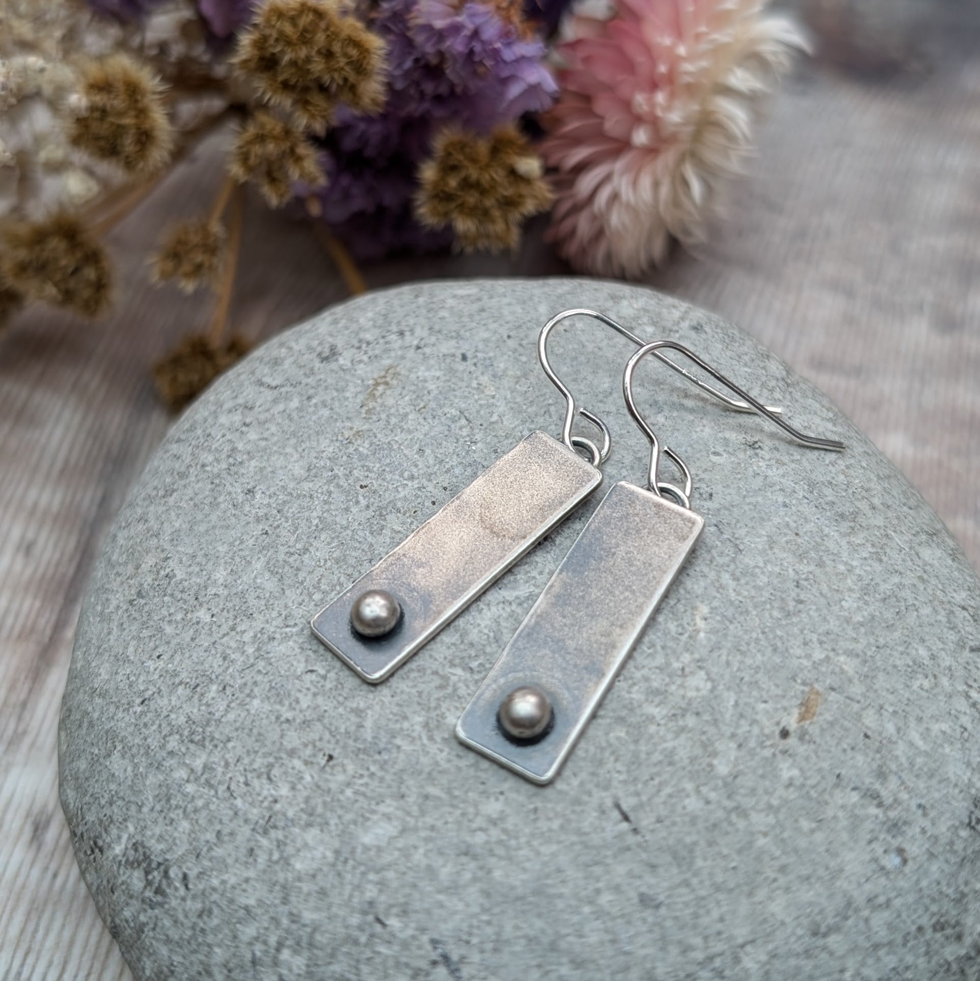 A pair of rectangular oxidized silver earrings with a small round silver pebble at the bottom of each rectangle, placed on a smooth gray stone.