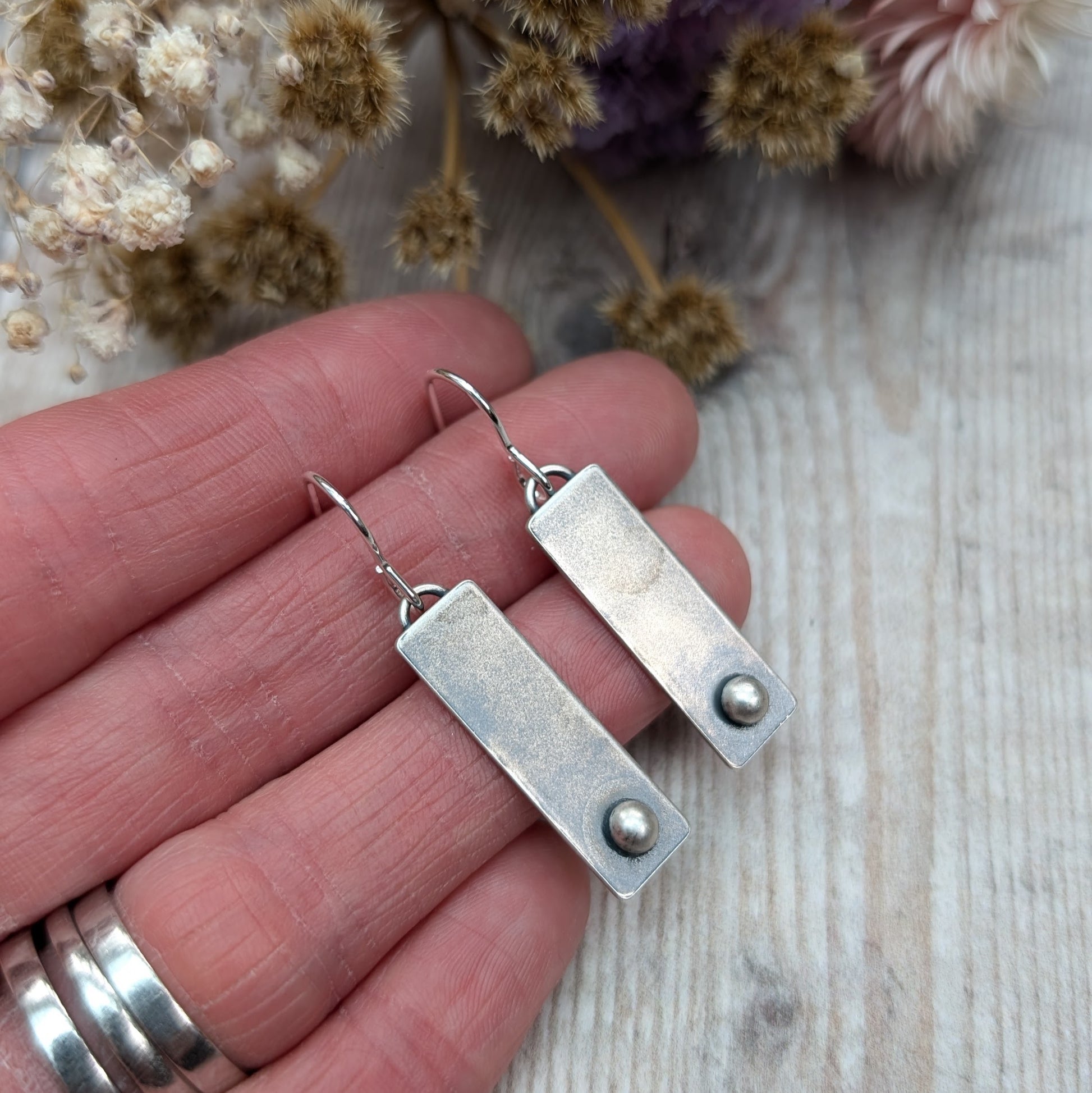 A pair of rectangular oxidized silver earrings with a small round silver pebble at the bottom of each rectangle, held in a person's hand for size reference, with a wooden background.