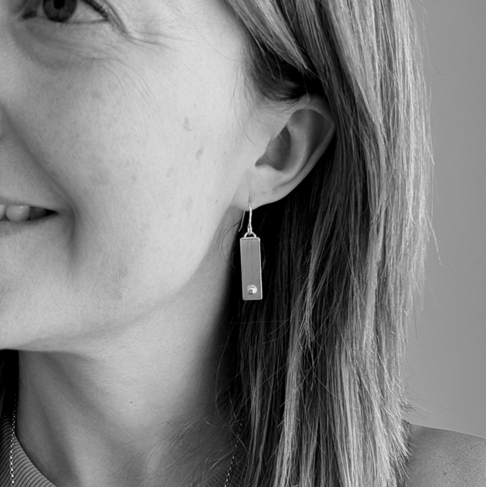 A pair of rectangular oxidized silver earrings with a small round silver pebble at the bottom of each rectangle, shown being worn for size reference.