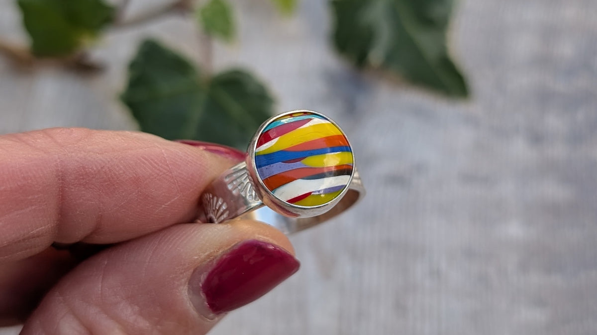 Close up view of Surfite Sunburst Ring held between fingers with green leaves in the background. 