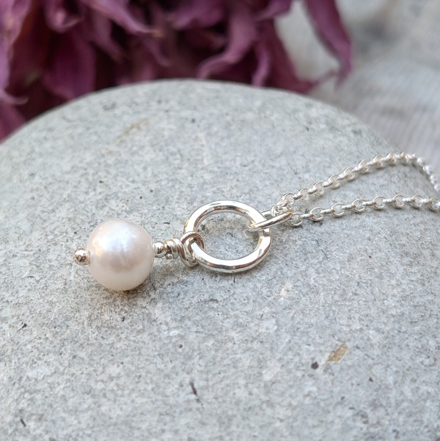 Close-up of a sterling silver necklace with a small silver circle and a single white pearl drop pendant, displayed on a smooth grey stone with dried flowers in the background.