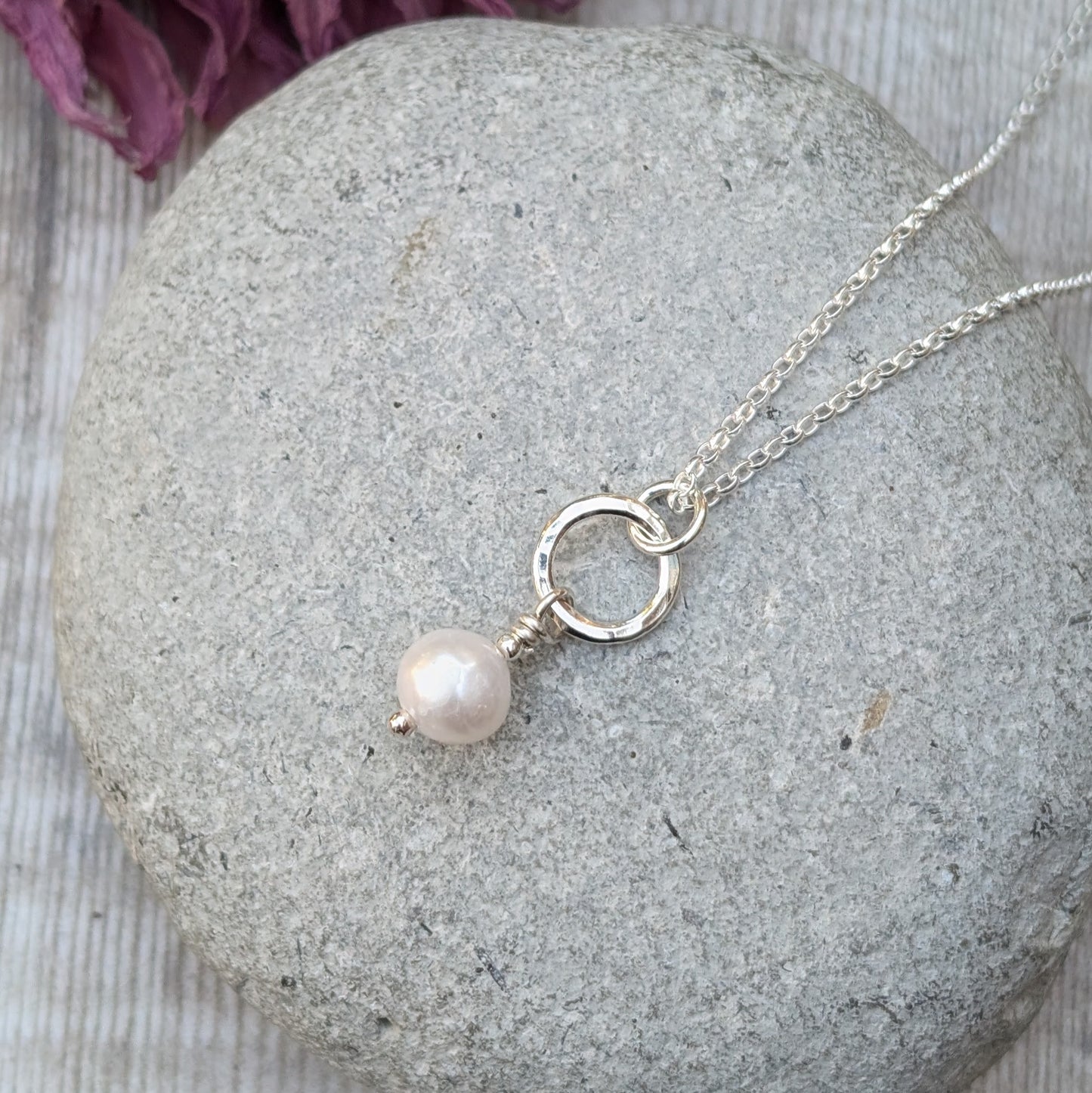 Close-up of a sterling silver necklace with a small silver circle and a single white pearl drop pendant, displayed on a smooth grey stone with dried flowers in the background.