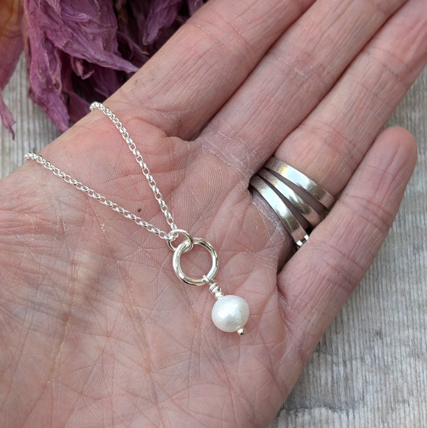 Close-up of a hand holding a delicate sterling silver necklace featuring a small silver circle with a single white pearl drop, with dried flowers in the background.