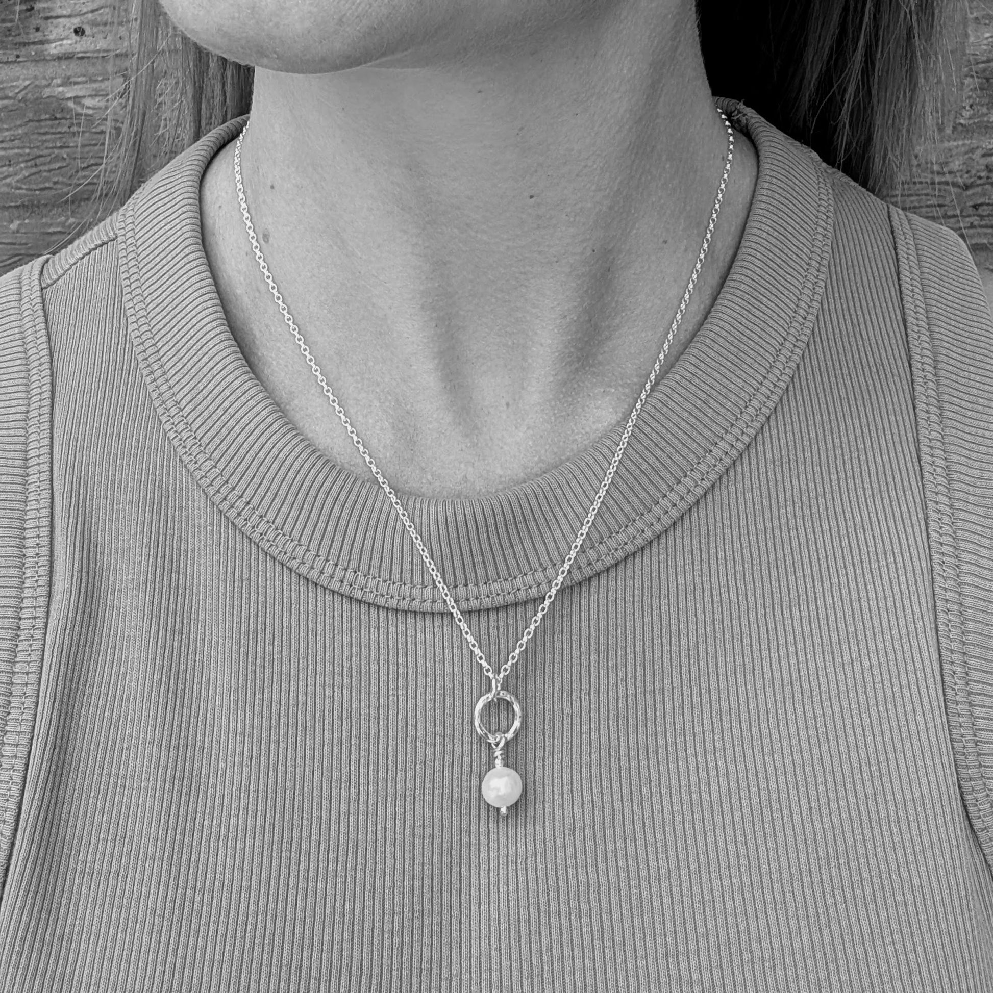 Black and white close-up of a person wearing a sterling silver necklace with a small silver circle and a white pearl drop pendant, resting just below the collarbone.