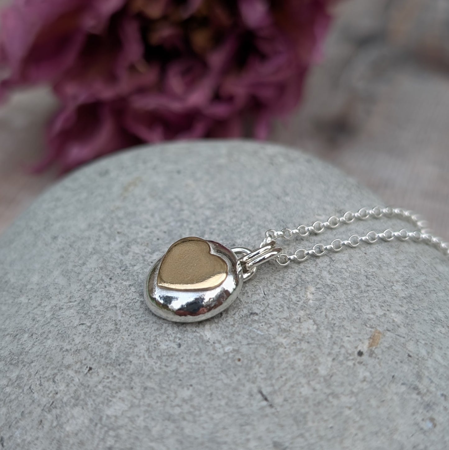 Recycled sterling silver pebble necklace with a 9ct recycled gold heart detail, placed on a smooth grey stone with a floral backdrop. The polished finish of the pendant reflects light beautifully