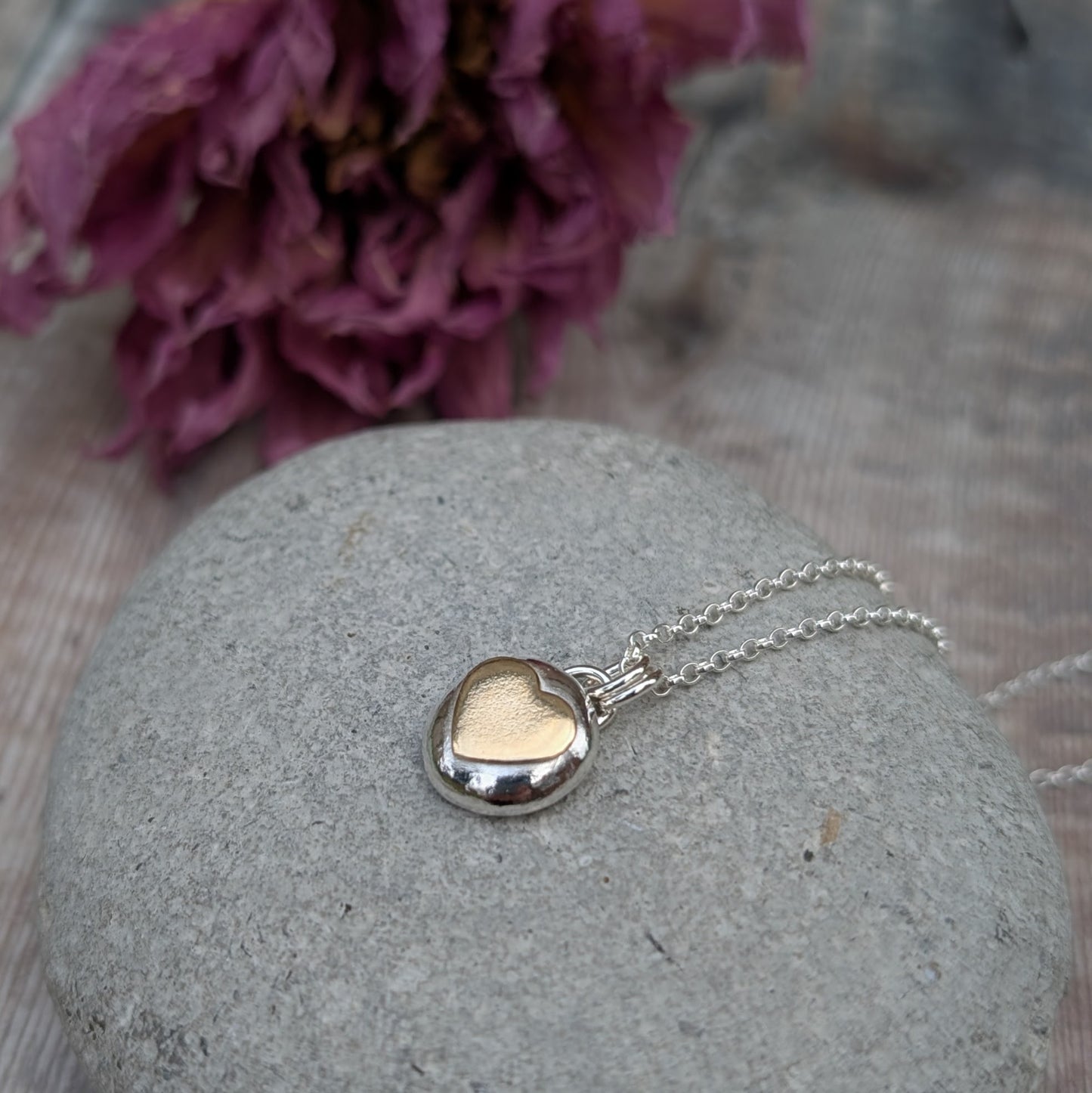 Recycled sterling silver pebble necklace with a 9ct recycled gold heart detail, placed on a smooth grey stone with a floral backdrop. The polished finish of the pendant reflects light beautifully