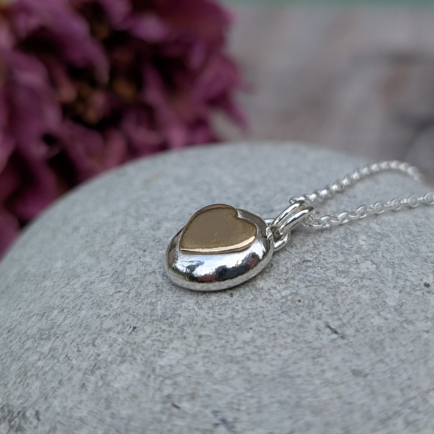 Recycled sterling silver pebble necklace with a 9ct recycled gold heart detail, placed on a smooth grey stone with a floral backdrop. The polished finish of the pendant reflects light beautifully