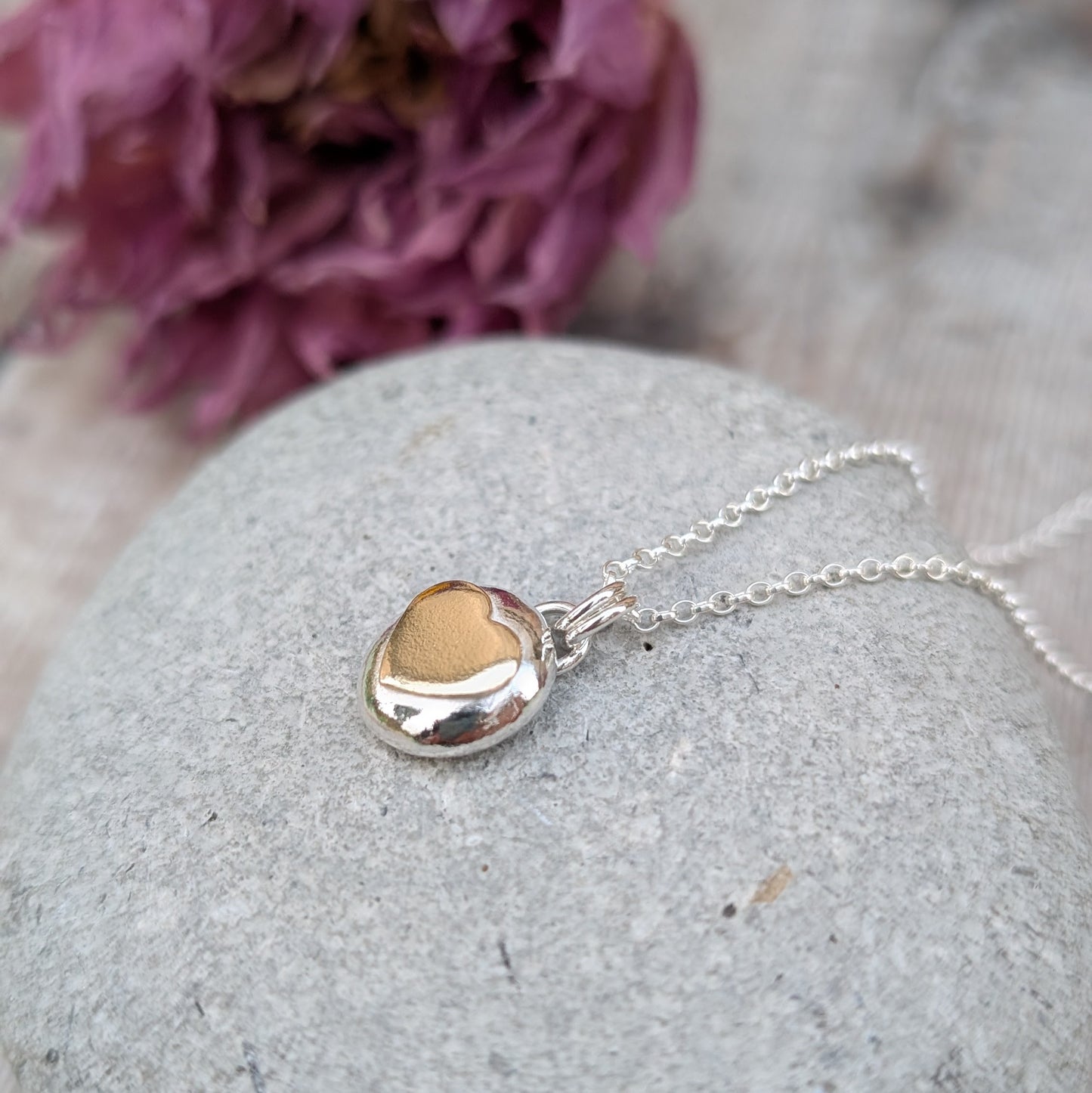 Recycled sterling silver pebble necklace with a 9ct recycled gold heart detail, placed on a smooth grey stone with a floral backdrop. The polished finish of the pendant reflects light beautifully