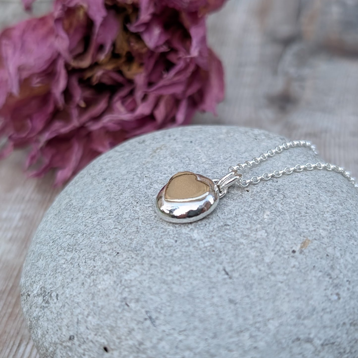 Recycled sterling silver pebble necklace with a 9ct recycled gold heart detail, placed on a smooth grey stone with a floral backdrop. The polished finish of the pendant reflects light beautifully