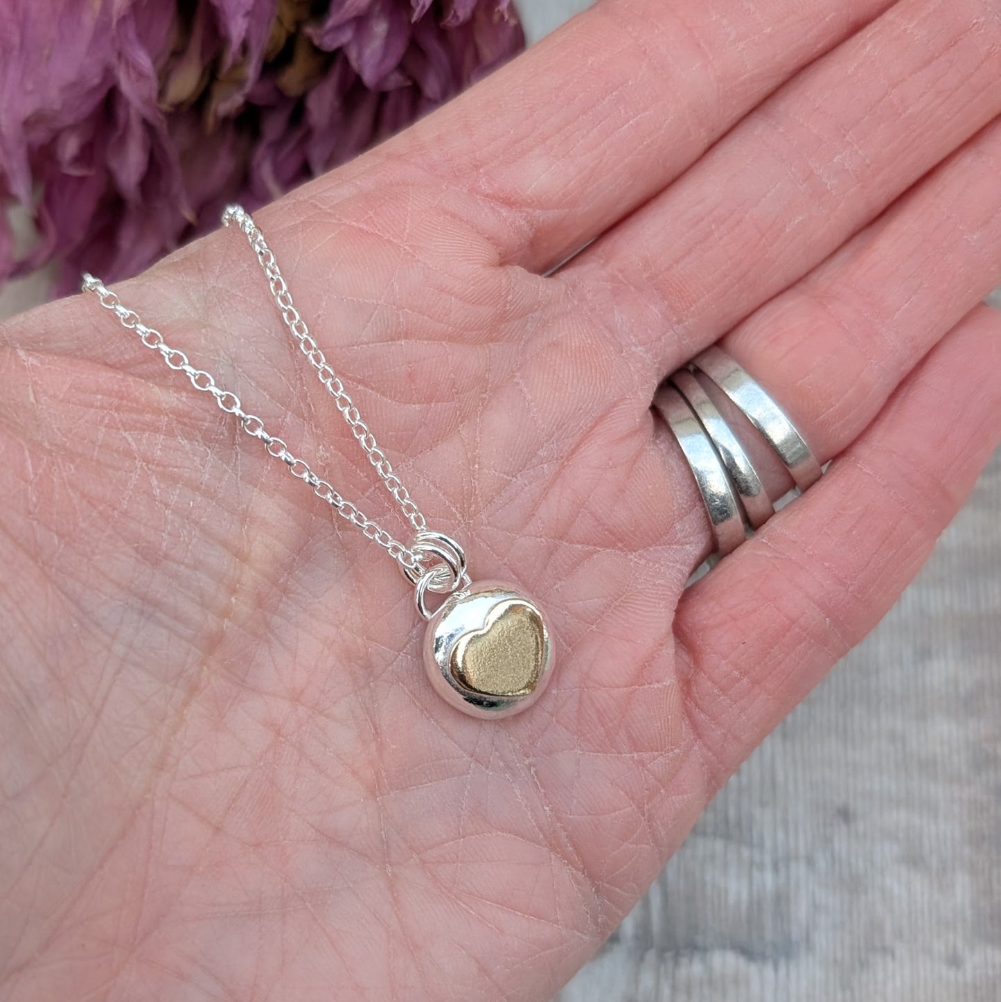 Recycled sterling silver pebble necklace with a 9ct recycled gold heart detail, shown resting on a hand for scale. The pendant has a smooth, polished finish and hangs on a delicate silver chain.