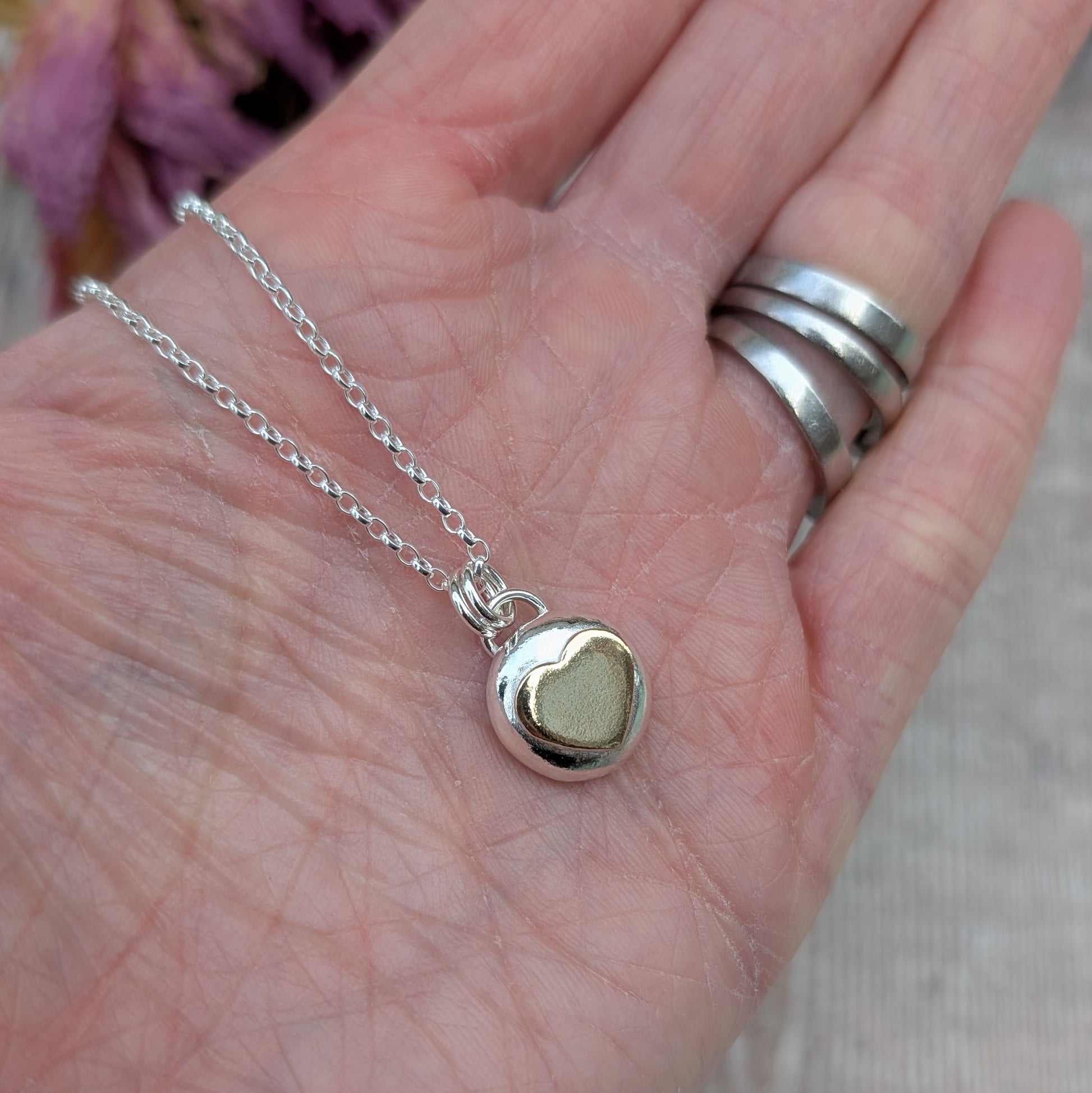 Recycled sterling silver pebble necklace with a 9ct recycled gold heart detail, shown resting on a hand for scale. The pendant has a smooth, polished finish and hangs on a delicate silver chain.