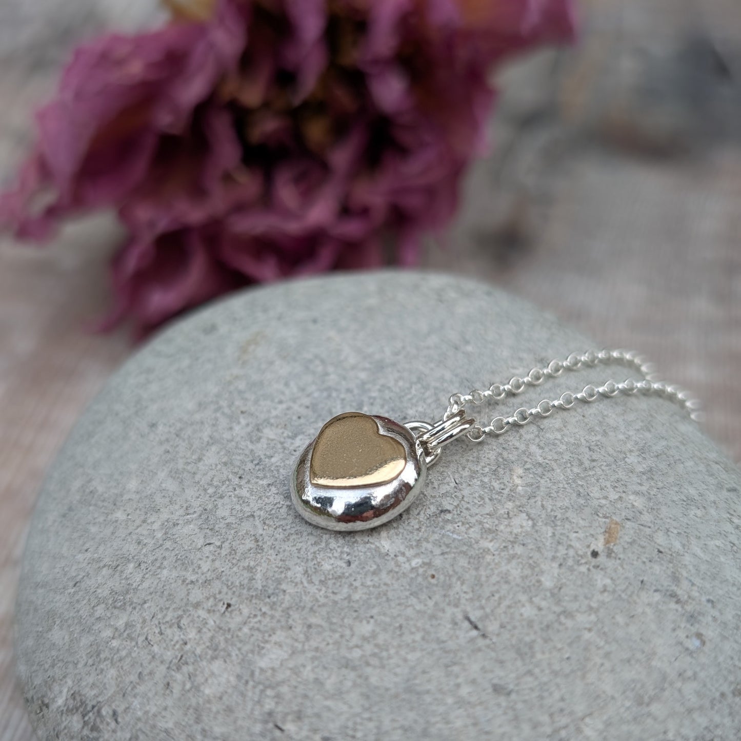 Recycled sterling silver pebble necklace with a 9ct recycled gold heart detail, placed on a smooth grey stone with a floral backdrop. The polished finish of the pendant reflects light beautifully