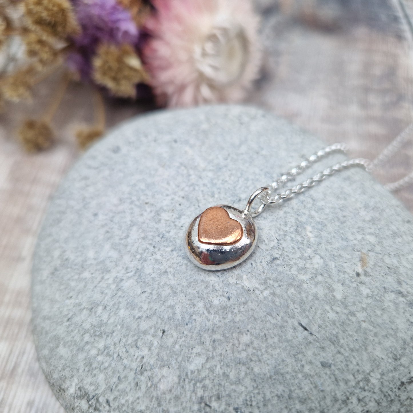 Sterling Silver round pebble shaped pendant with slightly raised silver heart shape at its centre. Suspended from silver chain via small circle link.