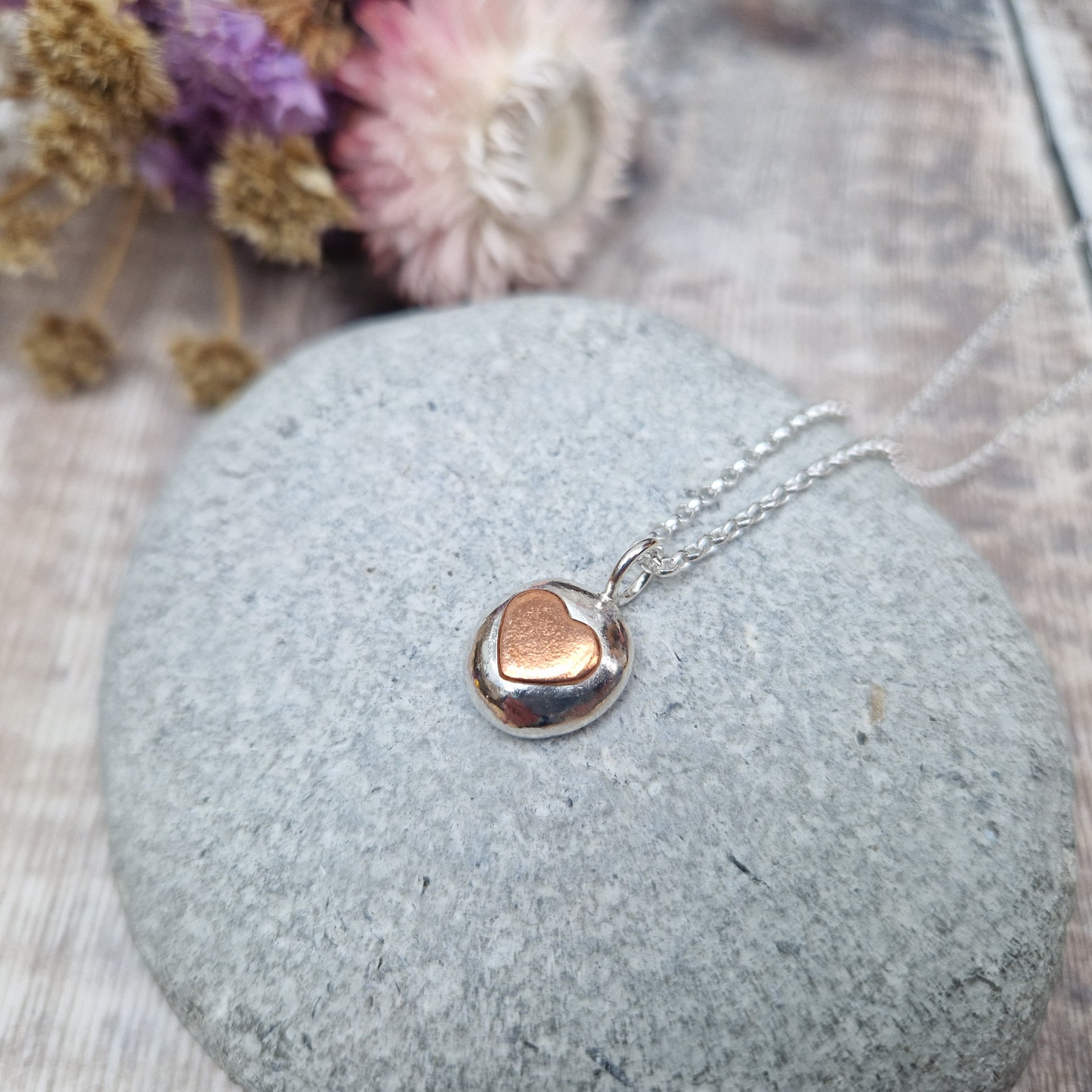 Sterling Silver round pebble shaped pendant with slightly raised silver heart shape at its centre. Suspended from silver chain via small circle link.