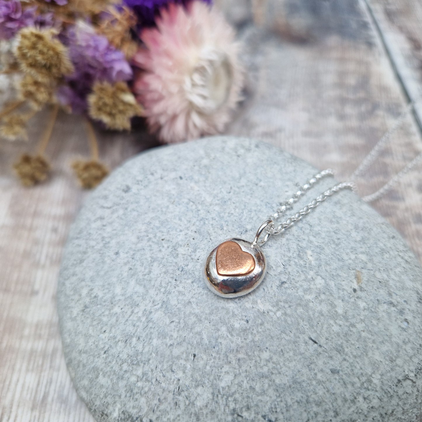 Sterling Silver round pebble shaped pendant with slightly raised silver heart shape at its centre. Suspended from silver chain via small circle link.