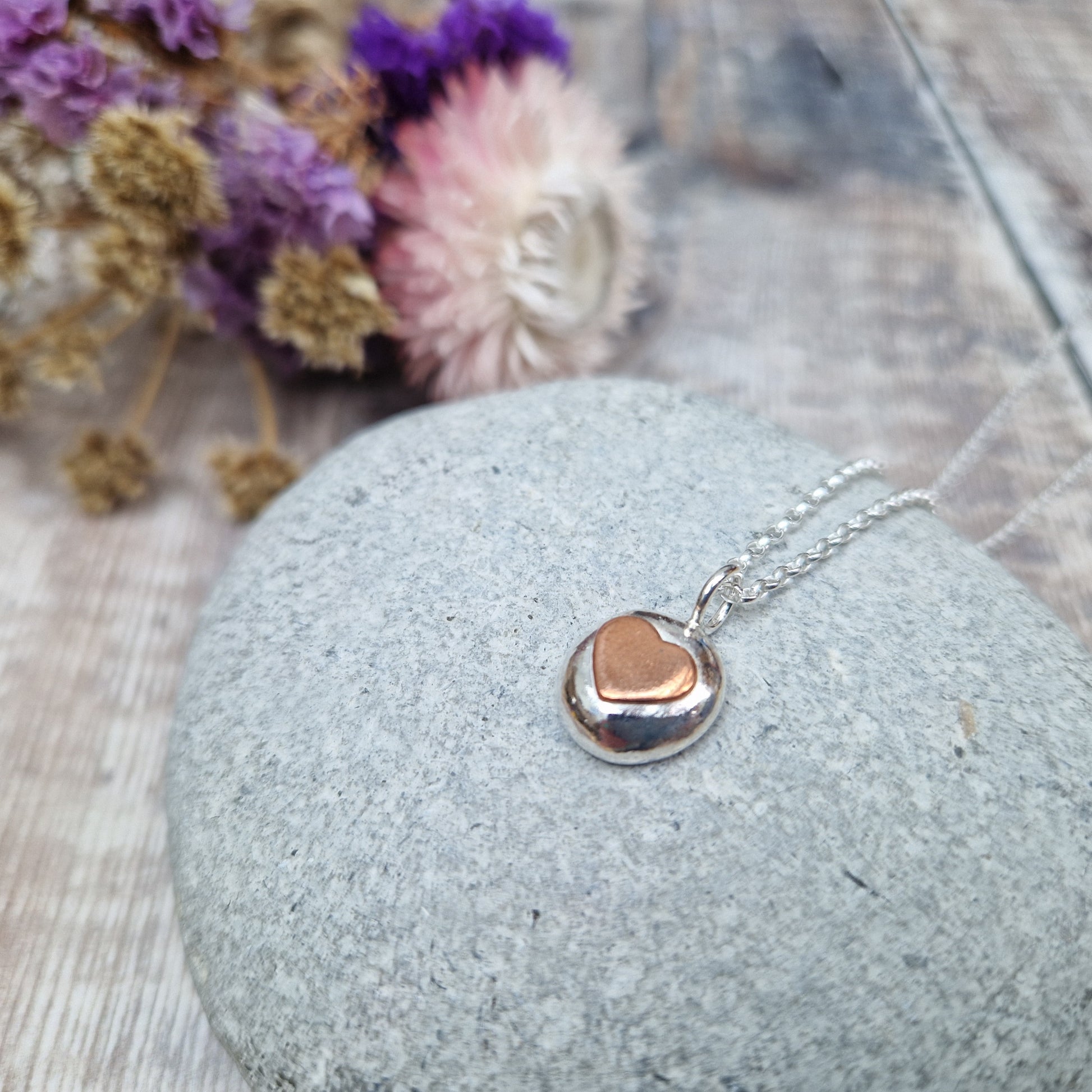 Sterling Silver round pebble shaped pendant with slightly raised silver heart shape at its centre. Suspended from silver chain via small circle link.