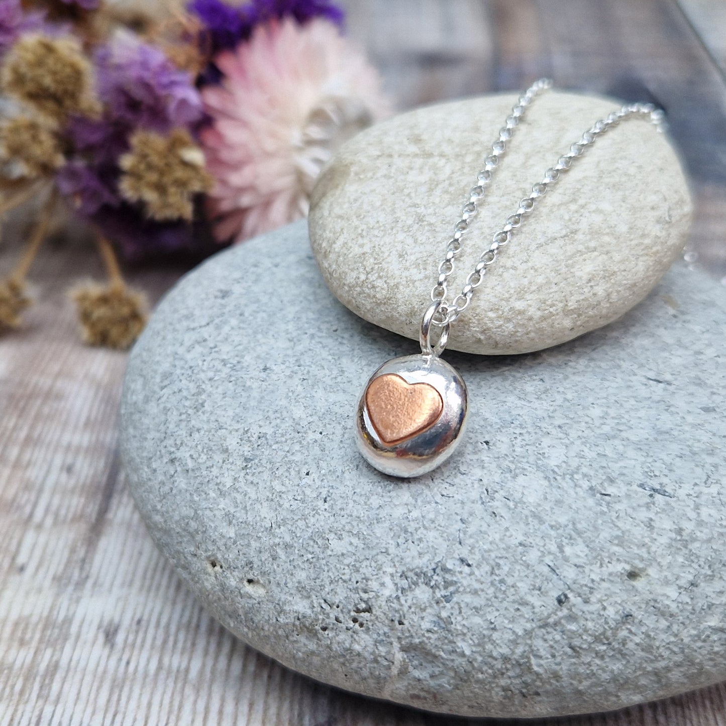 Sterling Silver round pebble shaped pendant with slightly raised silver heart shape at its centre. Suspended from silver chain via small circle link.