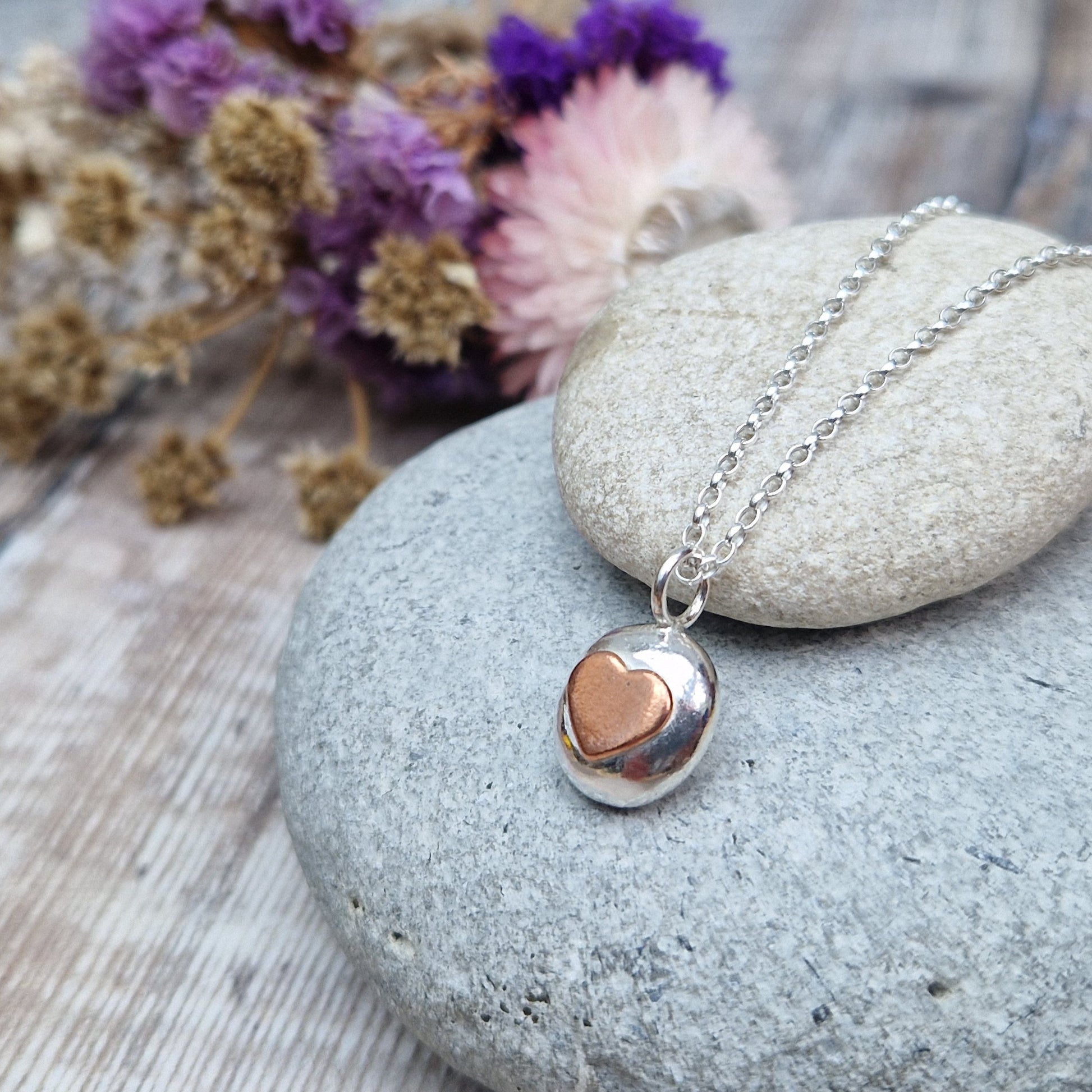 Sterling Silver round pebble shaped pendant with slightly raised silver heart shape at its centre. Suspended from silver chain via small circle link.