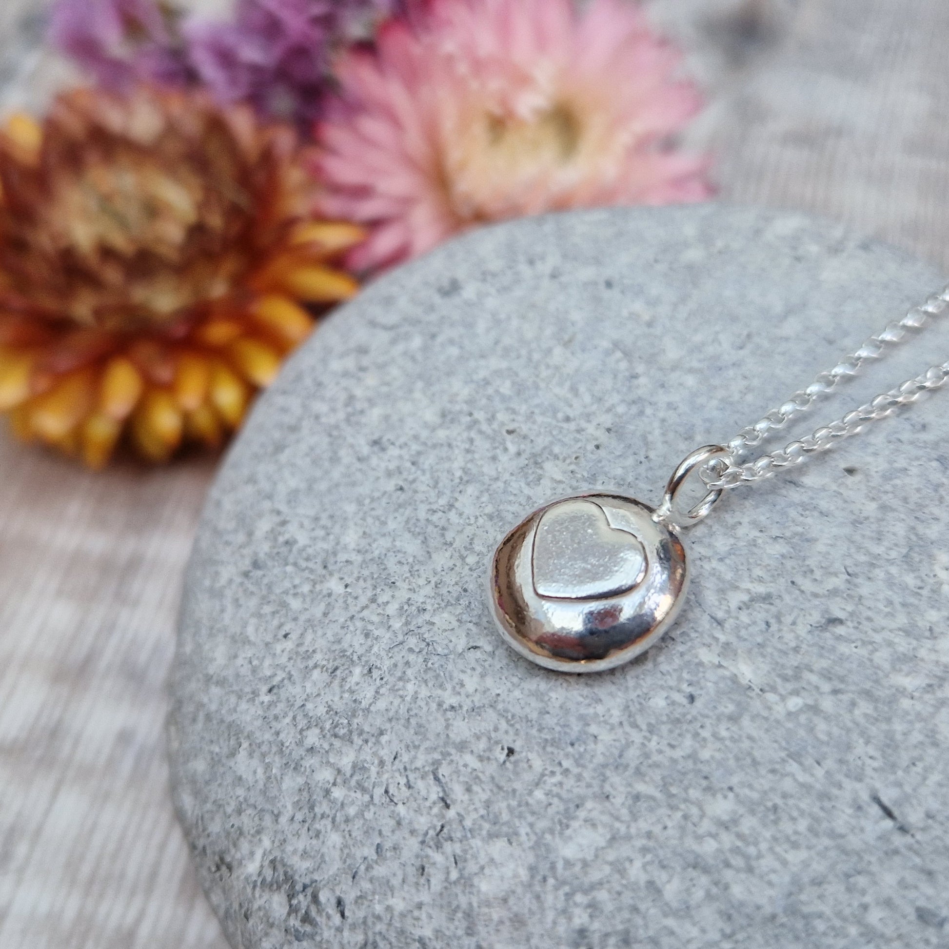 Sterling Silver round pebble shaped pendant with slightly raised silver heart shape at its centre. Suspended from silver chain via small circle link.