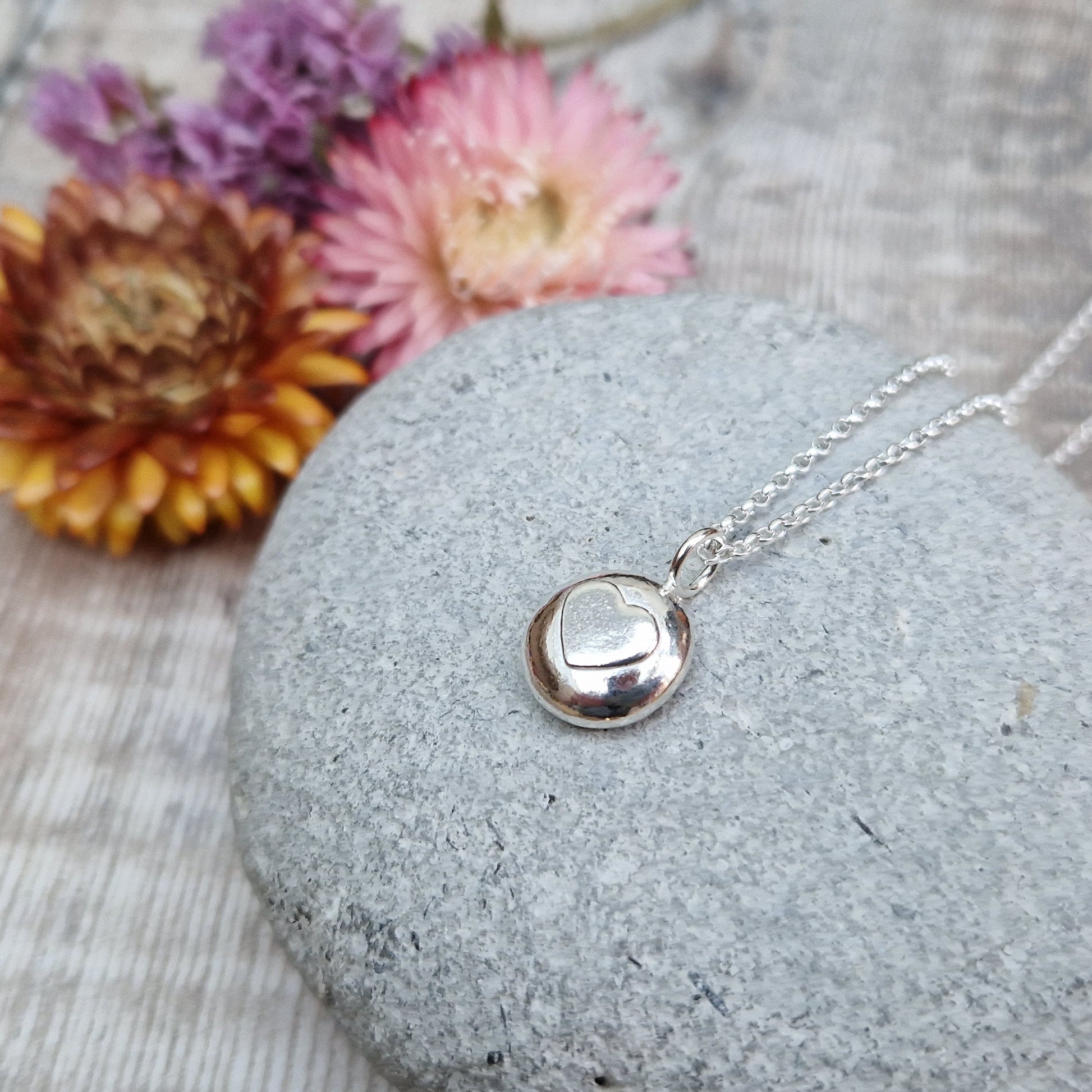 Sterling Silver round pebble shaped pendant with slightly raised silver heart shape at its centre. Suspended from silver chain via small circle link.