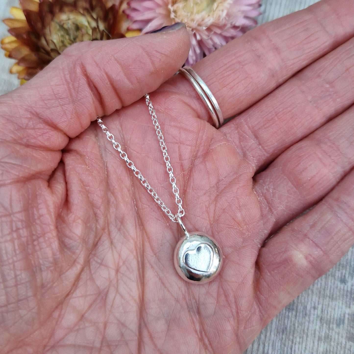 Sterling Silver round pebble shaped pendant with slightly raised silver heart shape at its centre. Suspended from silver chain via small circle link.