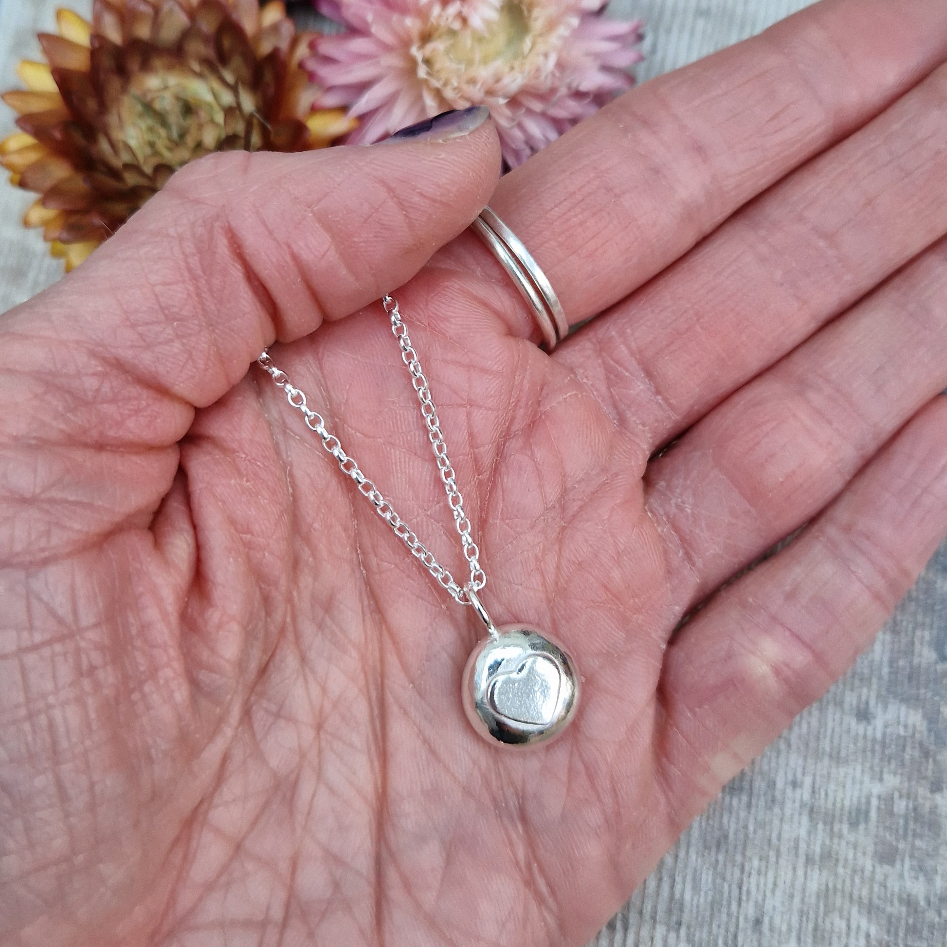 Sterling Silver round pebble shaped pendant with slightly raised silver heart shape at its centre. Suspended from silver chain via small circle link.