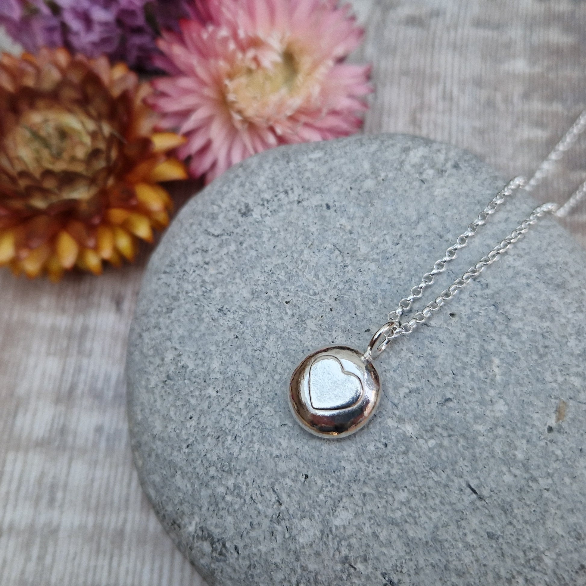 Sterling Silver round pebble shaped pendant with slightly raised silver heart shape at its centre. Suspended from silver chain via small circle link.