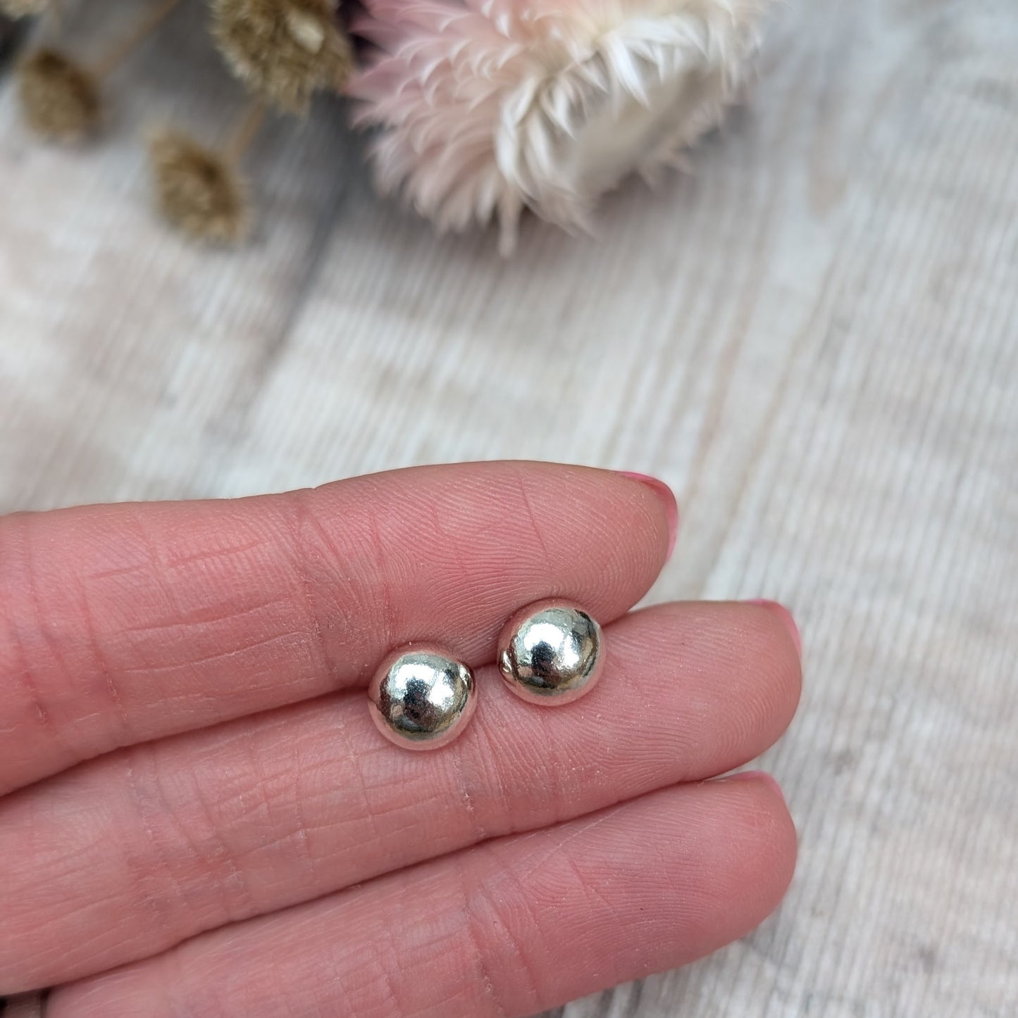 Close-up of a hand holding a pair of small, smooth, silver pebble stud earrings. The background features dried flowers on a wooden surface.