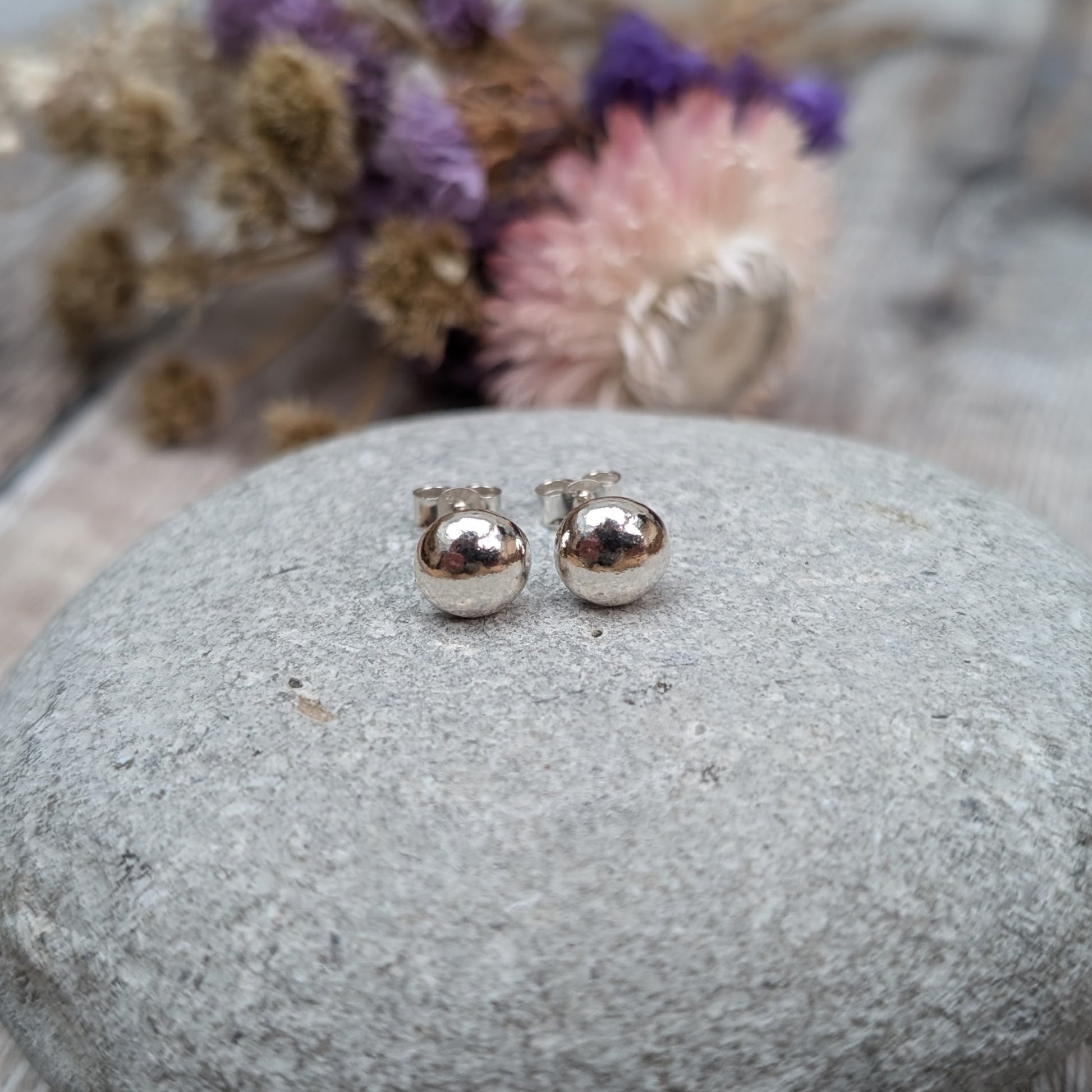 Pair of small, smooth, silver pebble stud earrings displayed on a grey stone. The background includes dried flowers with soft lighting.