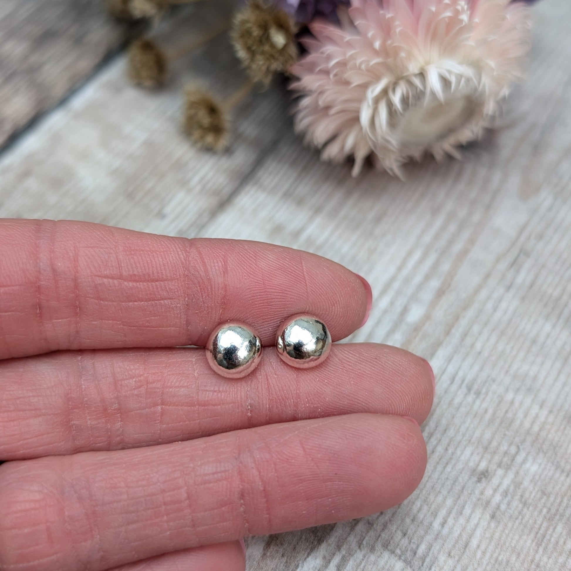 Close-up of a hand holding a pair of small, smooth, silver pebble stud earrings. The background features dried flowers on a wooden surface.