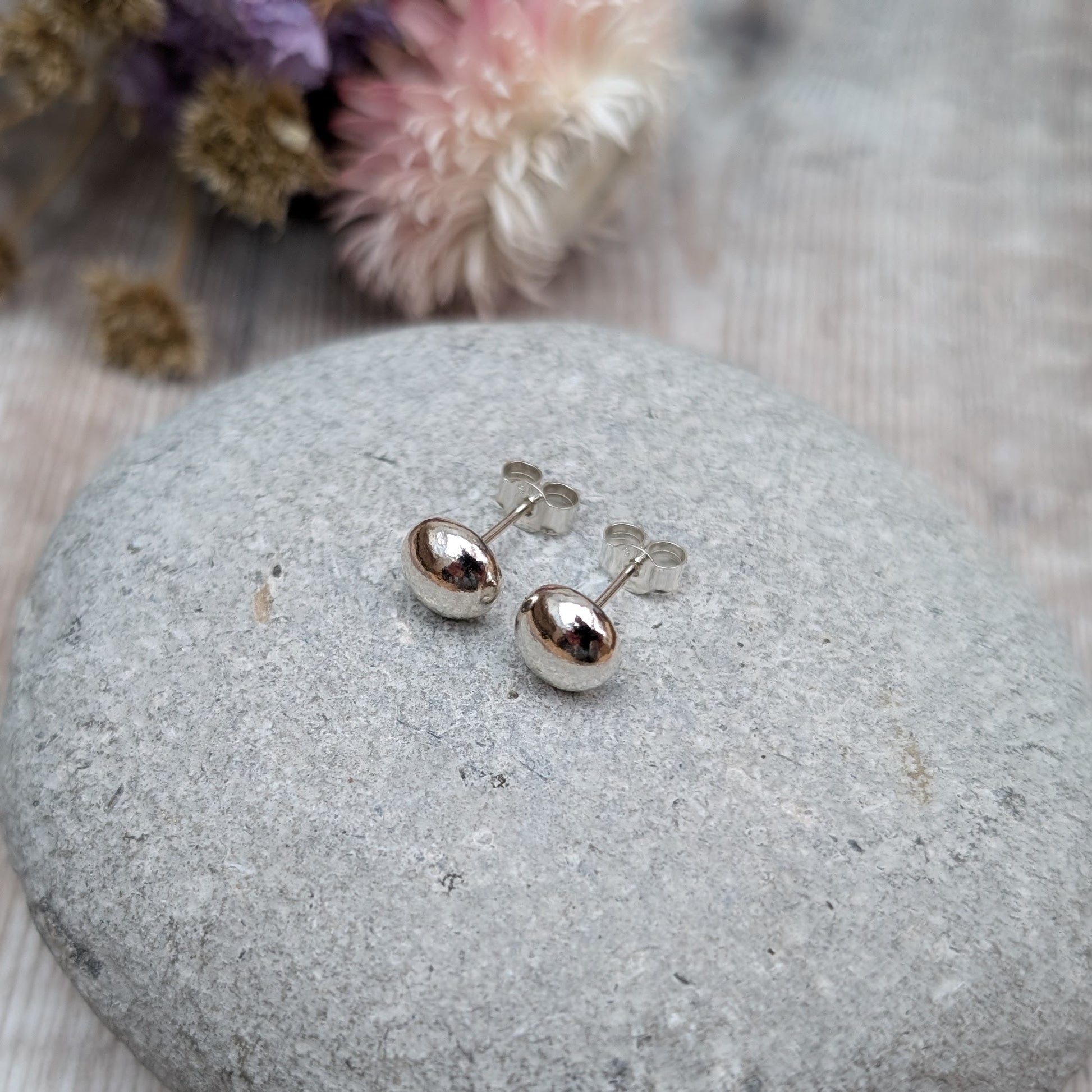 Pair of small, smooth, silver pebble stud earrings displayed on a grey stone. The background includes dried flowers with soft lighting.