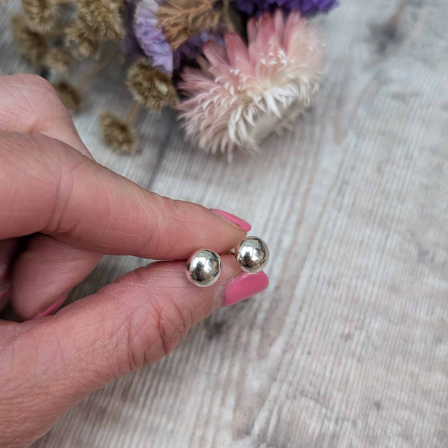 Close-up of a hand holding a pair of small, smooth, silver pebble stud earrings. The background features dried flowers on a wooden surface.