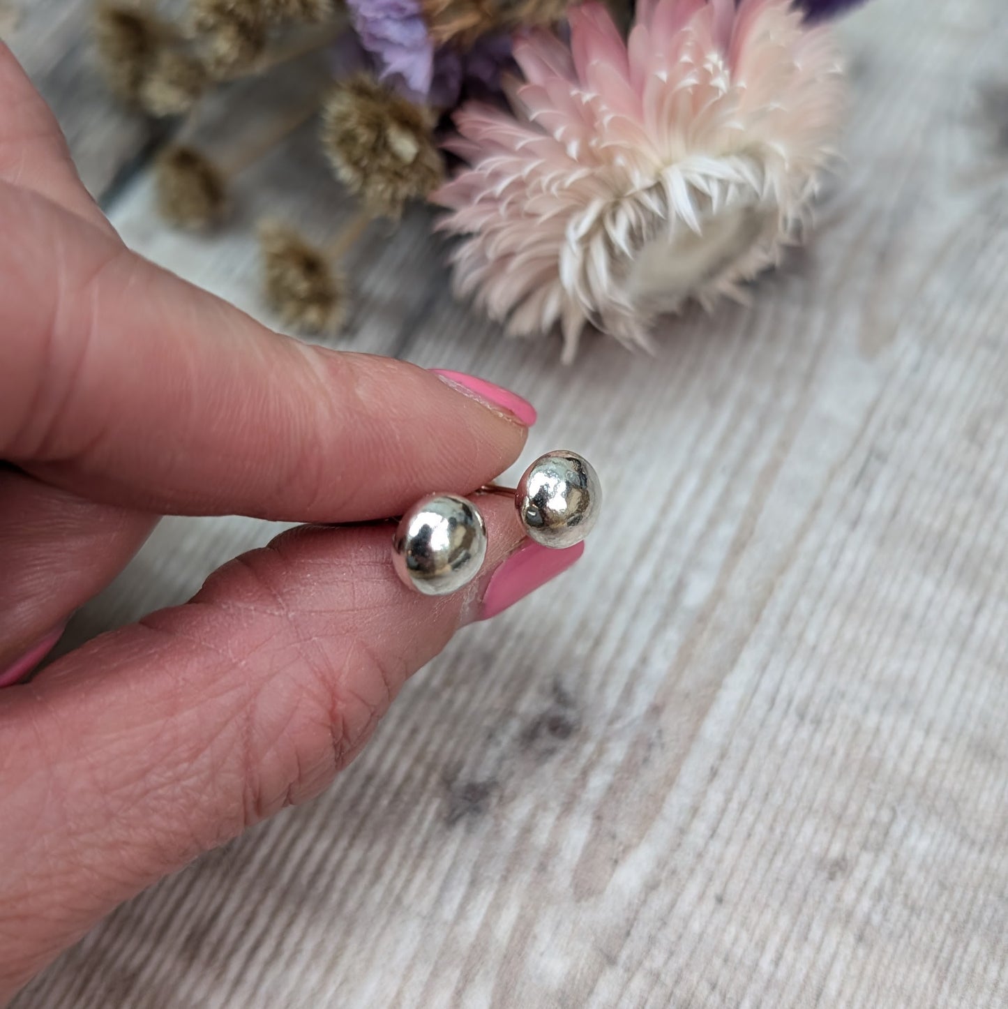 Close-up of a hand holding a pair of small, smooth, silver pebble stud earrings. The background features dried flowers on a wooden surface.