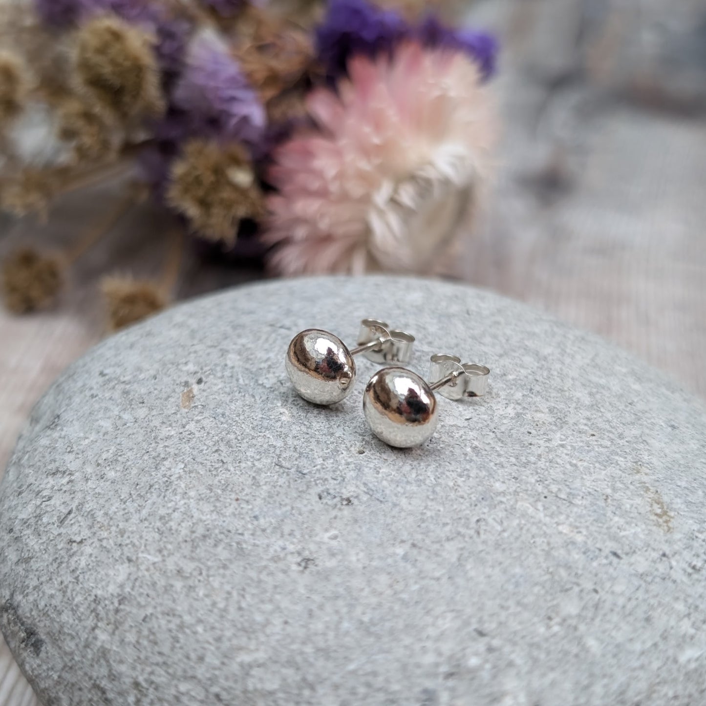 Pair of small, smooth, silver pebble stud earrings displayed on a grey stone. The background includes dried flowers with soft lighting.