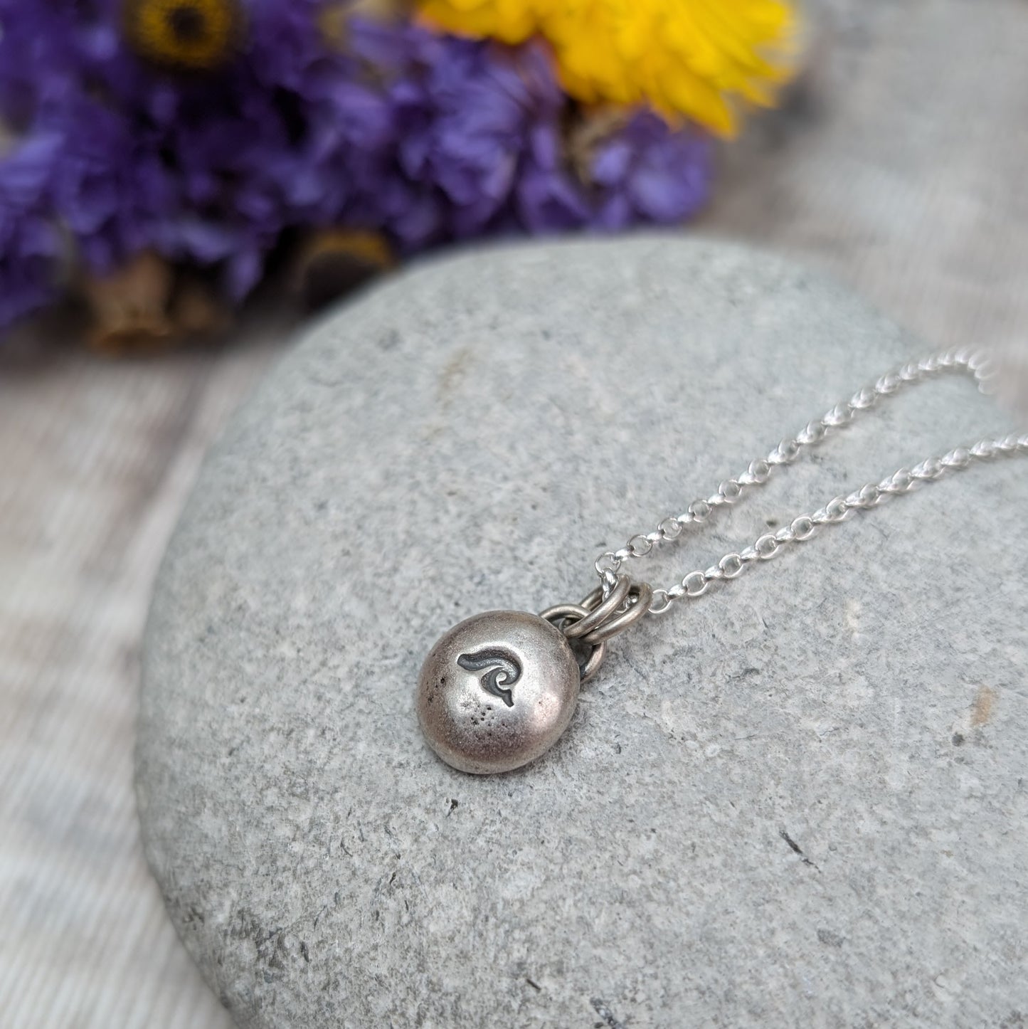 Oxidised Sterling Silver pebble shaped pendant, suspended from a silver chain via two small open circle links. A small wave shape is stamped on the smooth pebble surface.