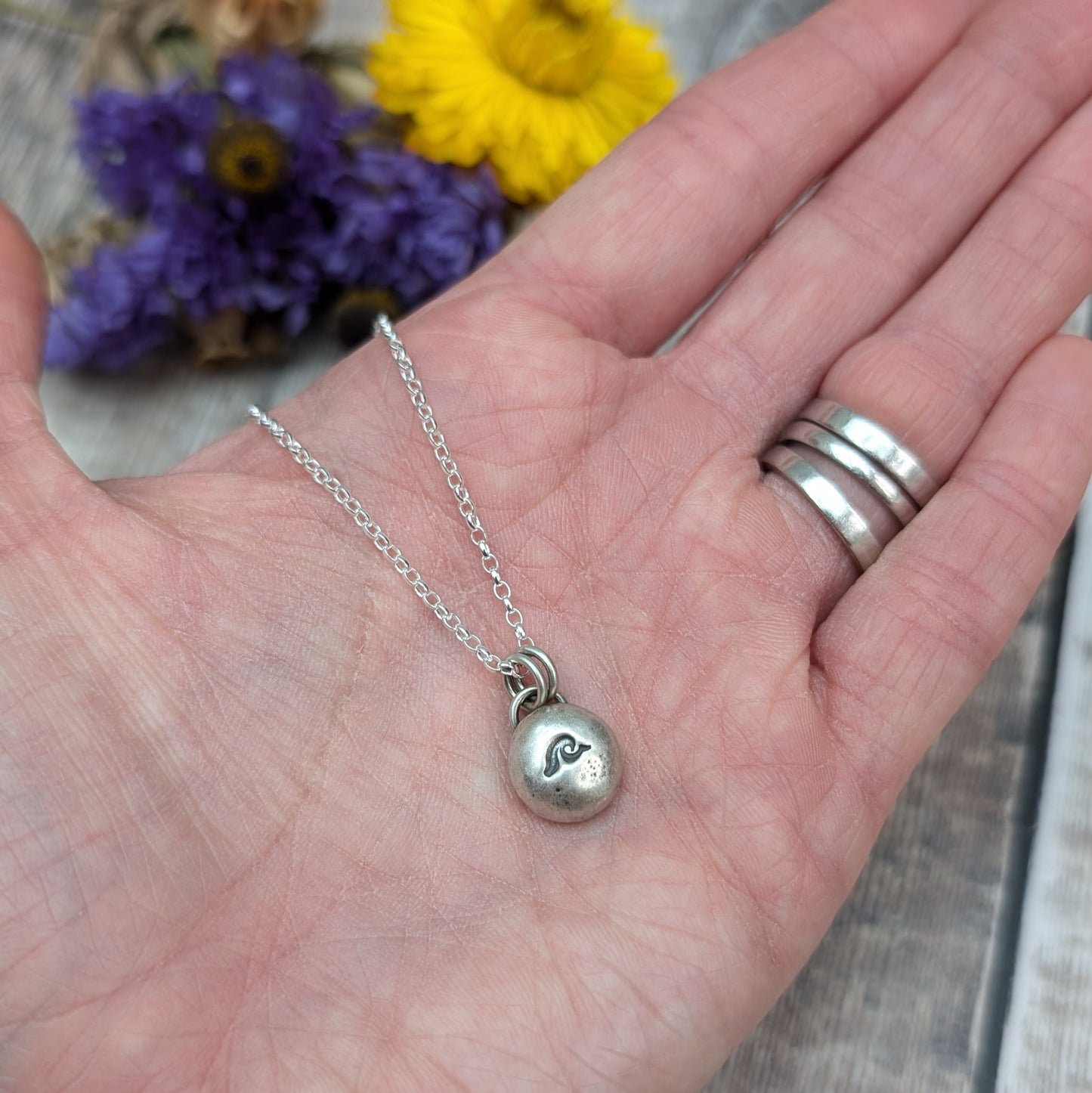 Oxidised Sterling Silver pebble shaped pendant, suspended from a silver chain via two small open circle links. A small wave shape is stamped on the smooth pebble surface.