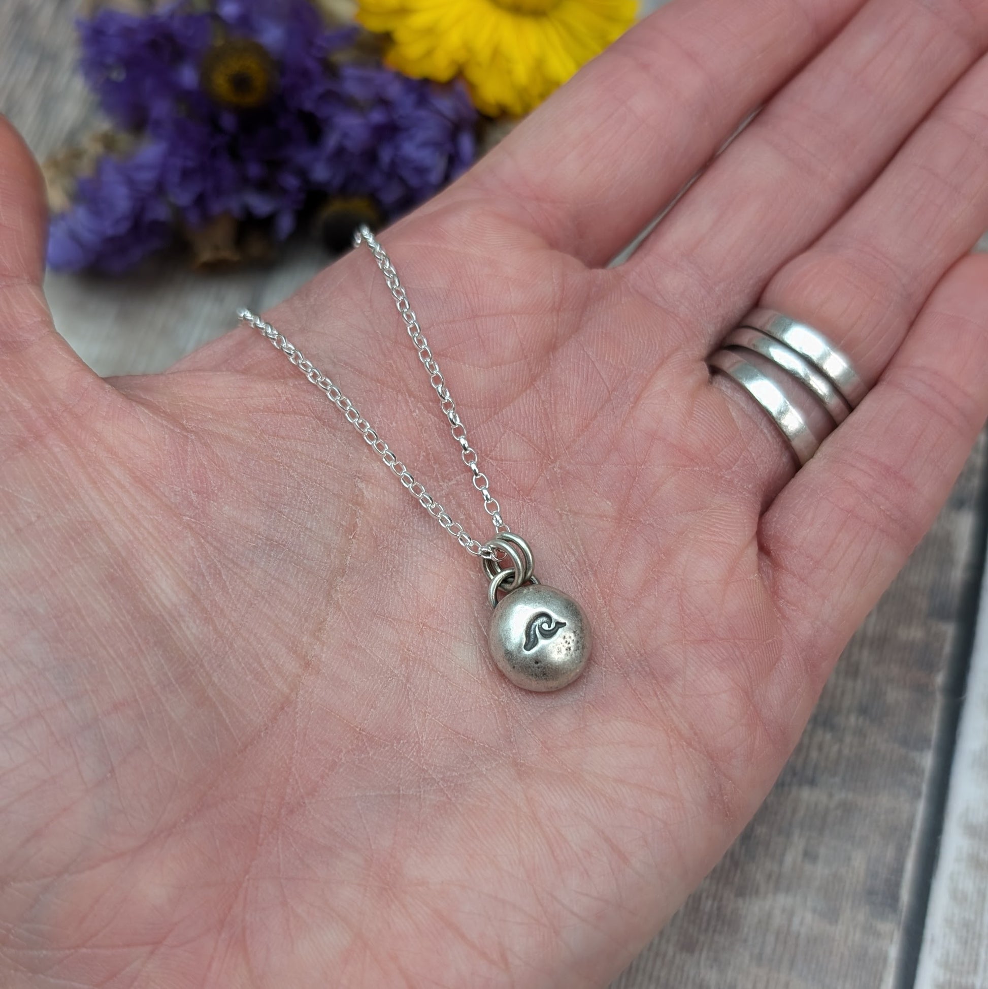 Oxidised Sterling Silver pebble shaped pendant, suspended from a silver chain via two small open circle links. A small wave shape is stamped on the smooth pebble surface.