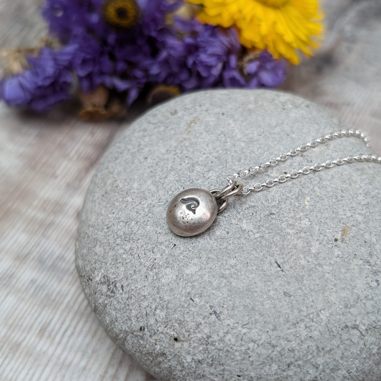 Oxidised Sterling Silver pebble shaped pendant, suspended from a silver chain via two small open circle links. A small wave shape is stamped on the smooth pebble surface.