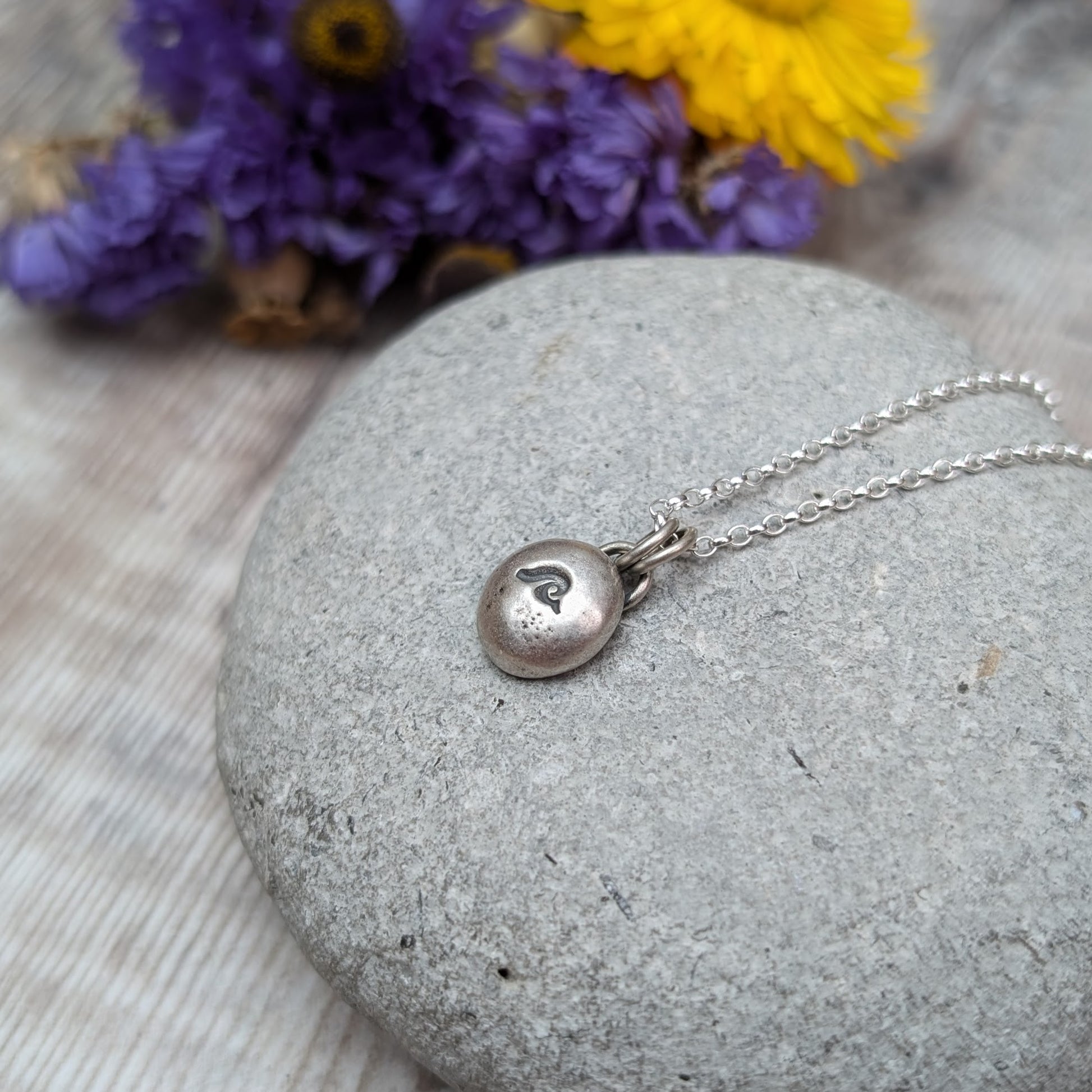 Oxidised Sterling Silver pebble shaped pendant, suspended from a silver chain via two small open circle links. A small wave shape is stamped on the smooth pebble surface.