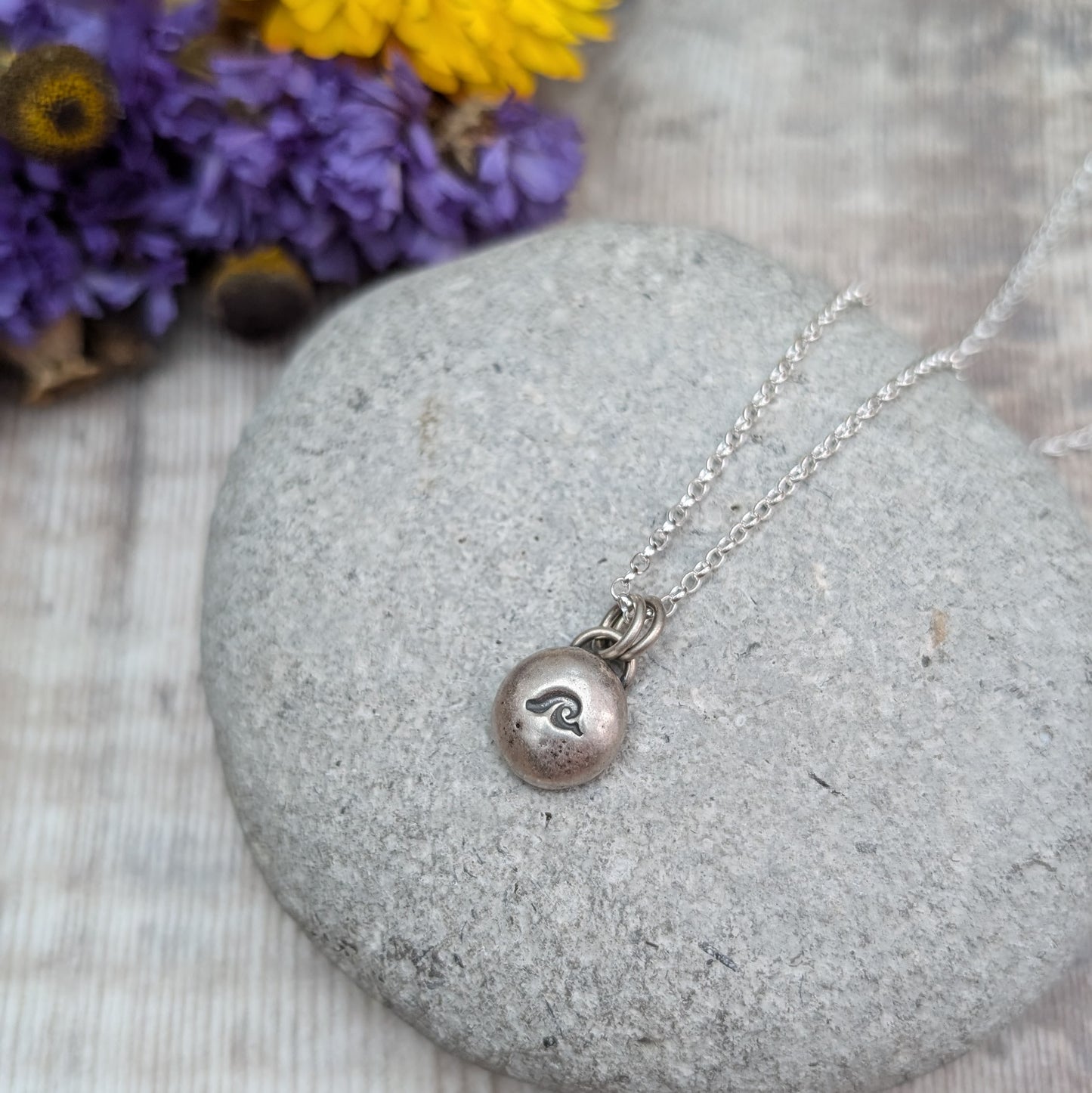 Oxidised Sterling Silver pebble shaped pendant, suspended from a silver chain via two small open circle links. A small wave shape is stamped on the smooth pebble surface.