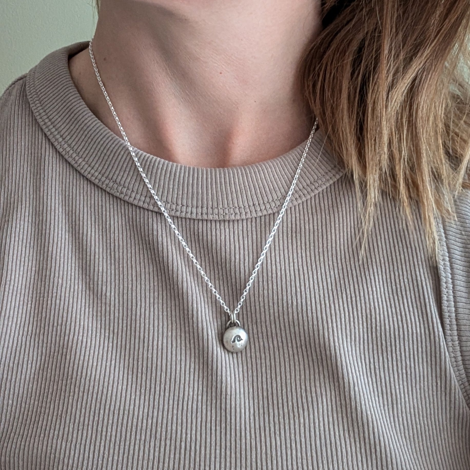 Oxidised Sterling Silver pebble shaped pendant, suspended from a silver chain via two small open circle links. A small wave shape is stamped on the smooth pebble surface.