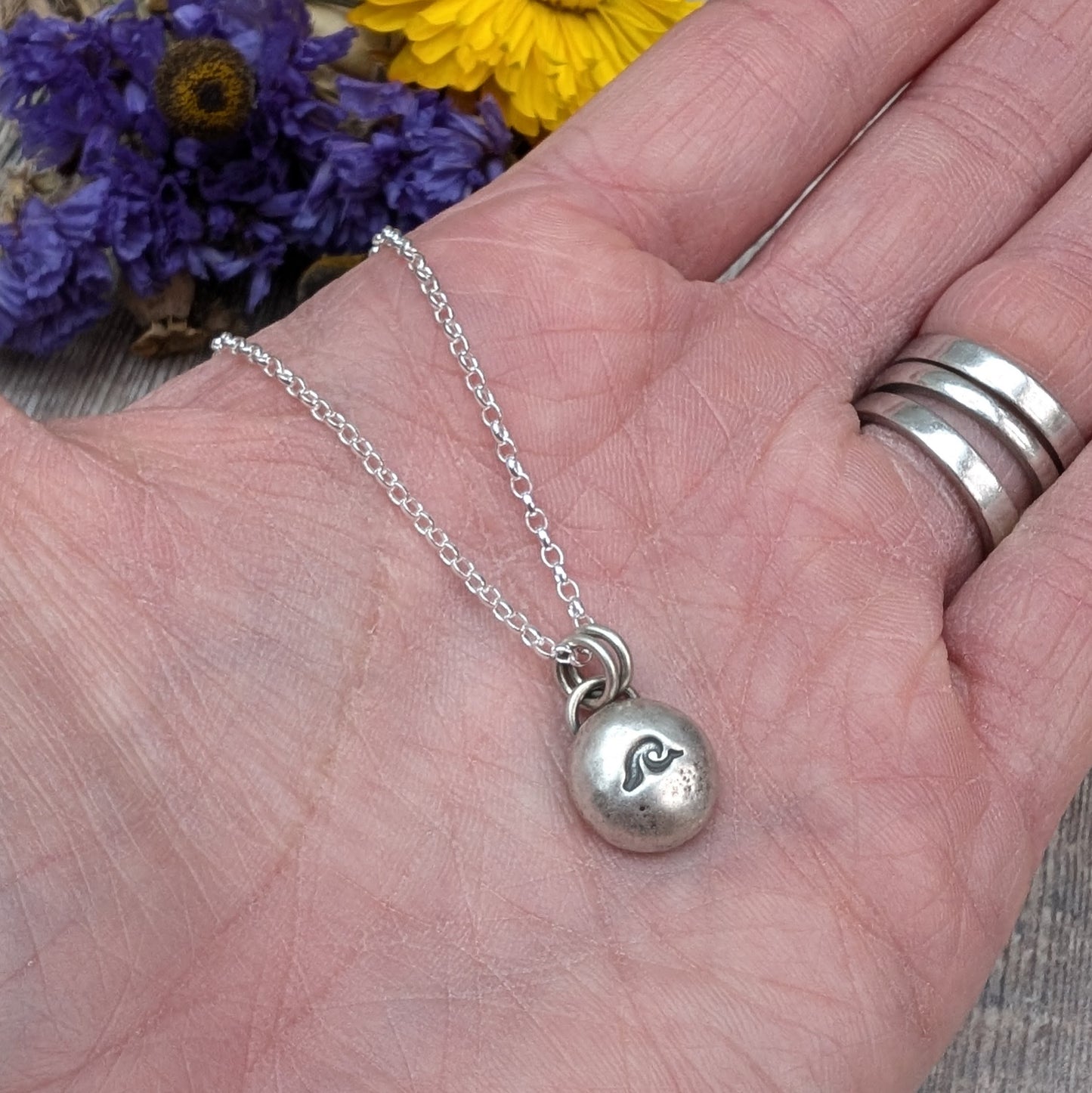 Oxidised Sterling Silver pebble shaped pendant, suspended from a silver chain via two small open circle links. A small wave shape is stamped on the smooth pebble surface.