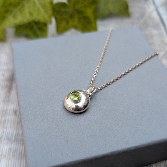 Peridot pebble necklace photographed on a grey jewellery box, featuring a handmade recycled Sterling Silver pebble pendant with a sparkling green Peridot gemstone.