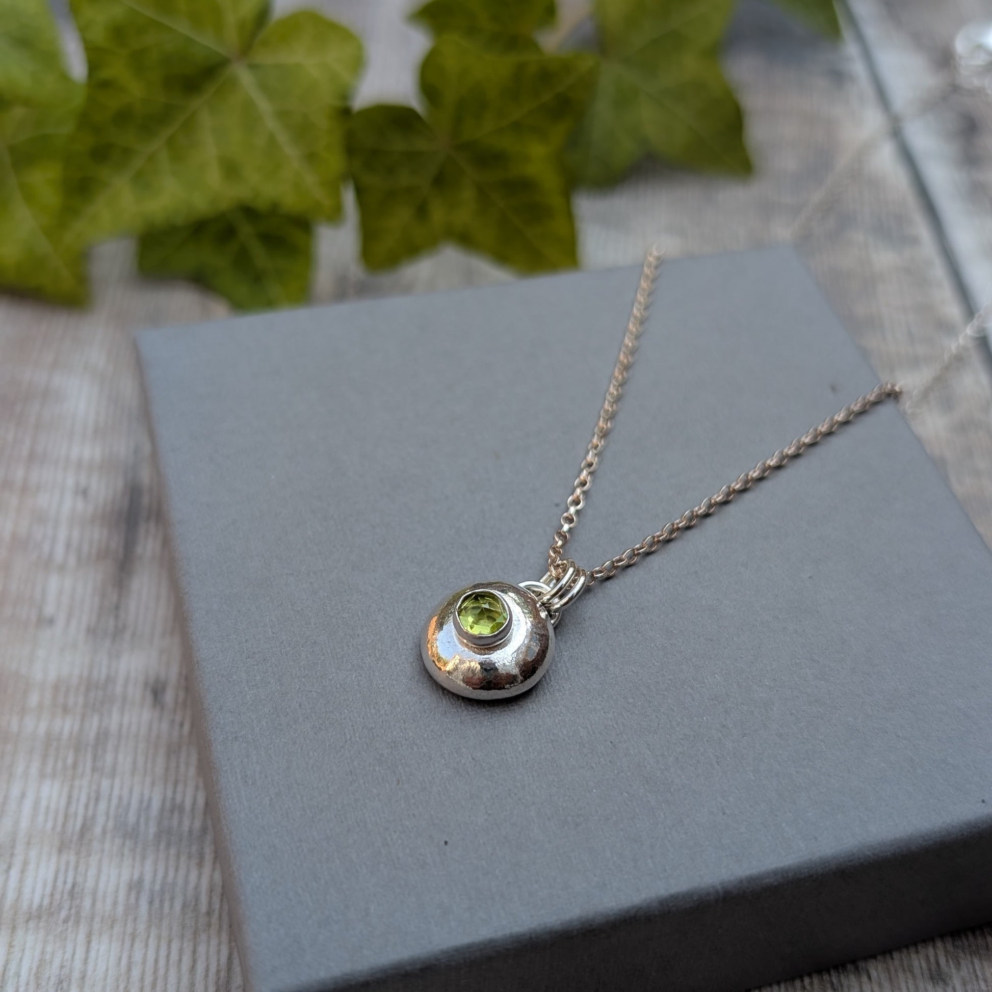 Peridot pebble necklace displayed on a grey jewellery box, showing a handmade recycled Sterling Silver pebble pendant with a green Peridot birthstone.