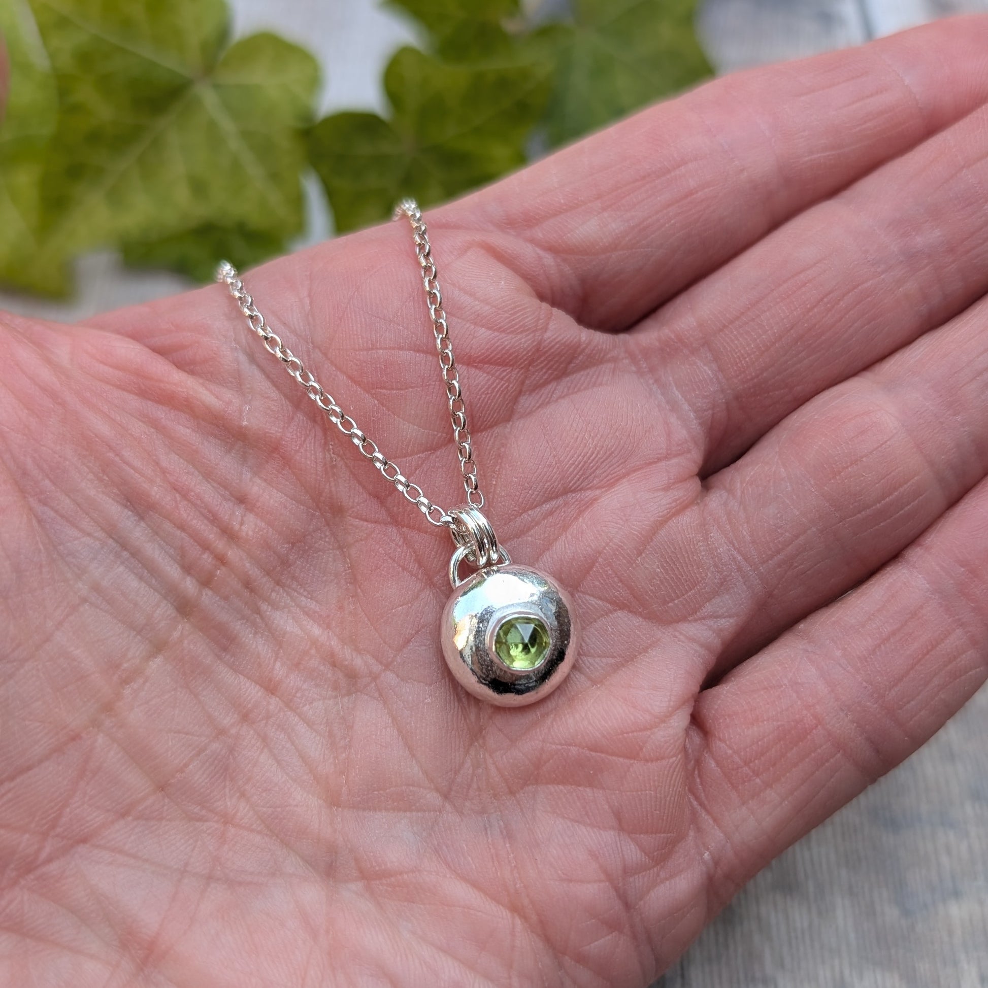 Close up of a peridot pebble necklace in the hand, featuring a polished recycled Sterling Silver pebble pendant with a bright green Peridot birthstone.