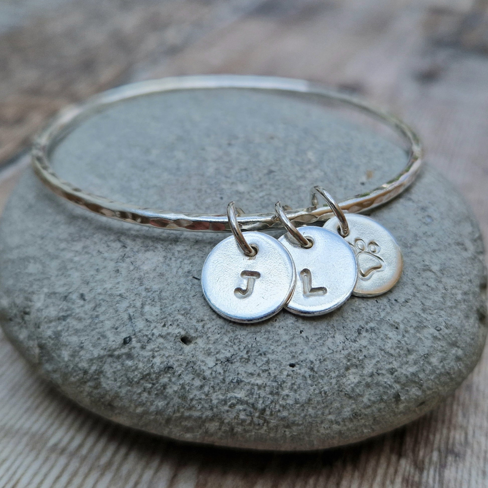 Hammered texture silver bangle decorated with three small disc pendants each suspended from the bangle via small open circle links. Example initials and a paw shaped stamp have been added to each disc.