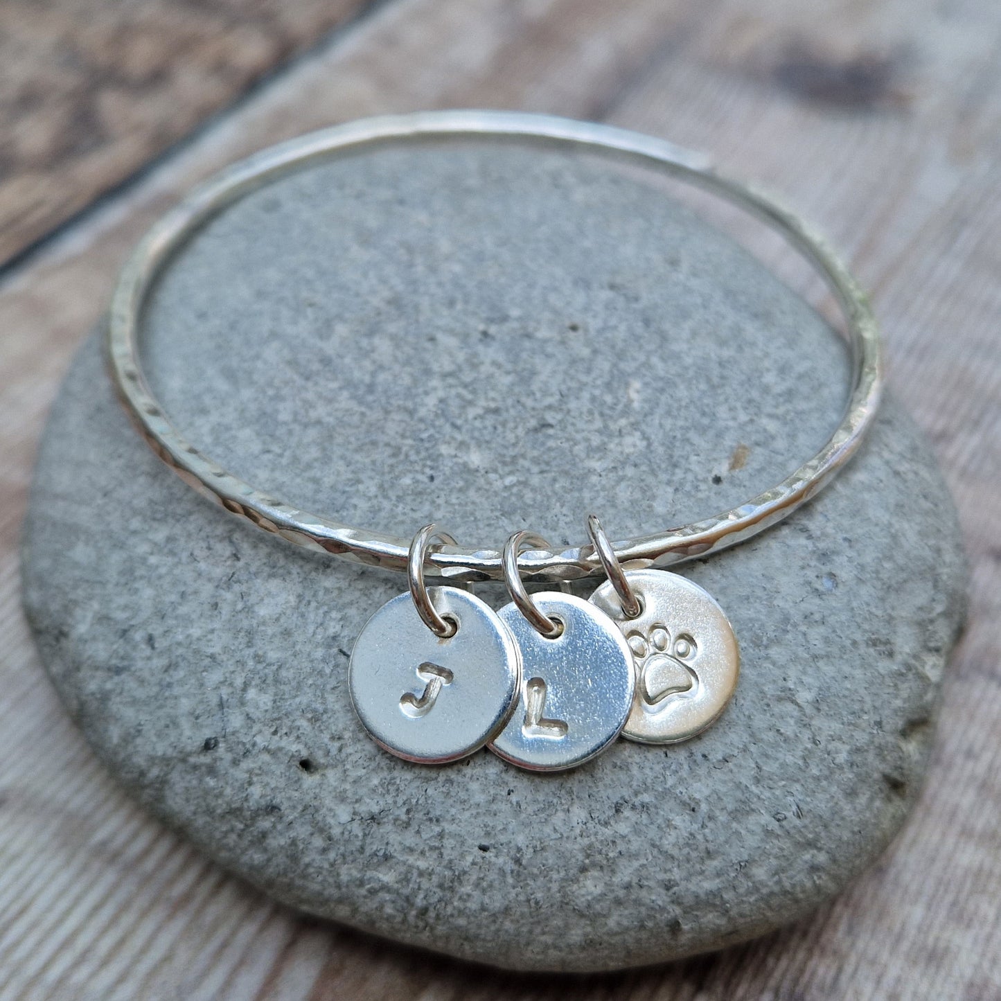 Hammered texture silver bangle decorated with three small disc pendants each suspended from the bangle via small open circle links. Example initials and a paw shaped stamp have been added to each disc.