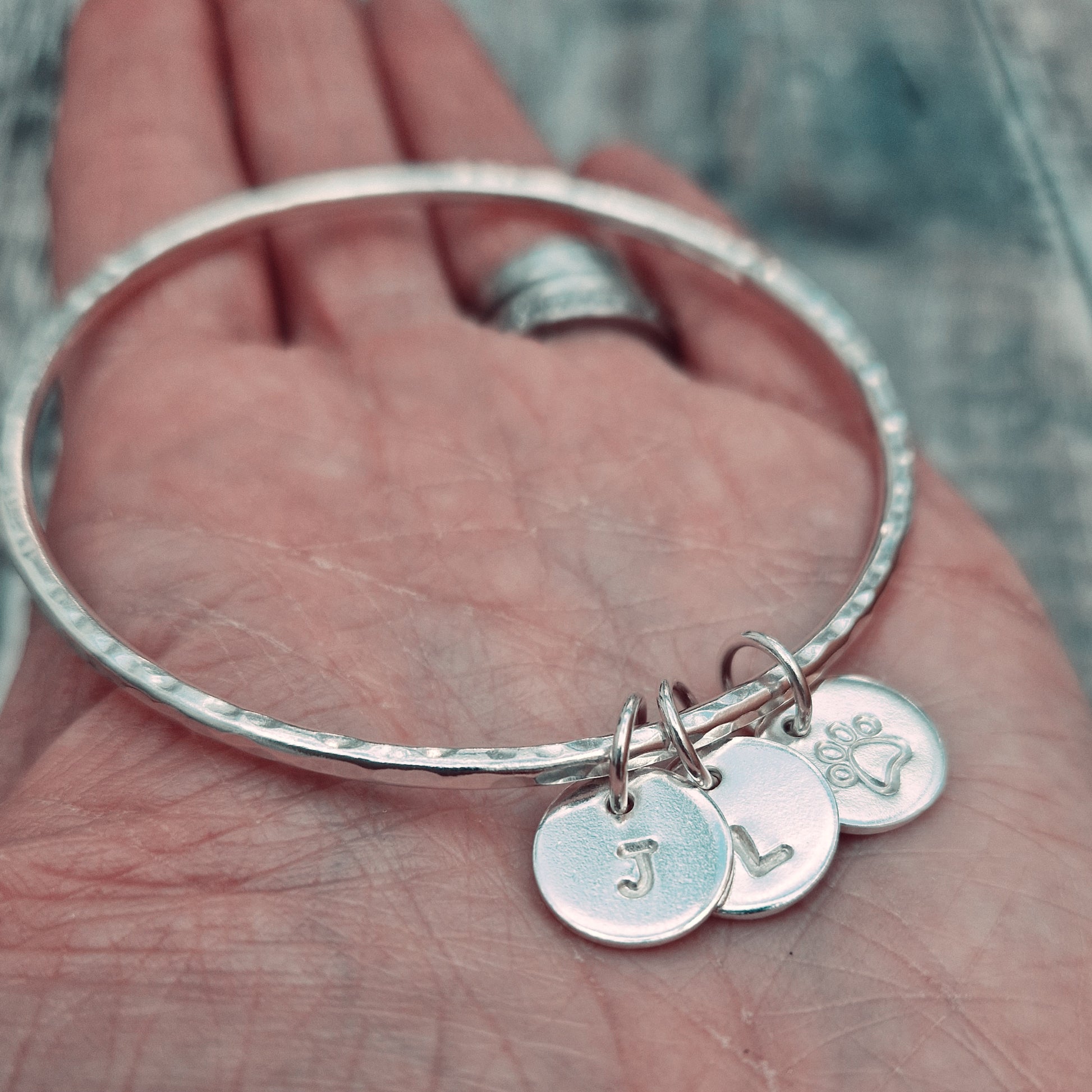 Hammered texture silver bangle decorated with three small disc pendants each suspended from the bangle via small open circle links. Example initials and a paw shaped stamp have been added to each disc.