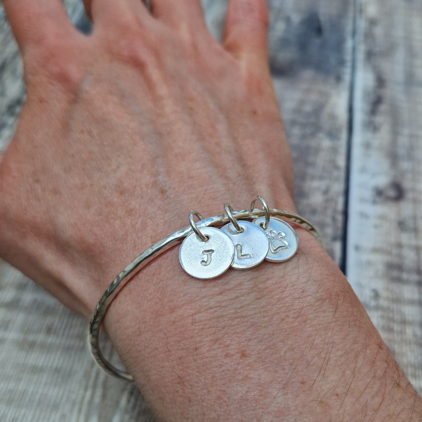 Hammered texture silver bangle decorated with three small disc pendants each suspended from the bangle via small open circle links. Example initials and a paw shaped stamp have been added to each disc.